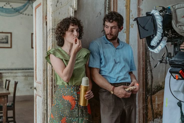 A woman applies lipstick while holding a thermos, standing next to a man with bread in his hand. They are on a film set, with a camera and crew equipment visible to the right. The setting appears vintage with worn walls and furniture.