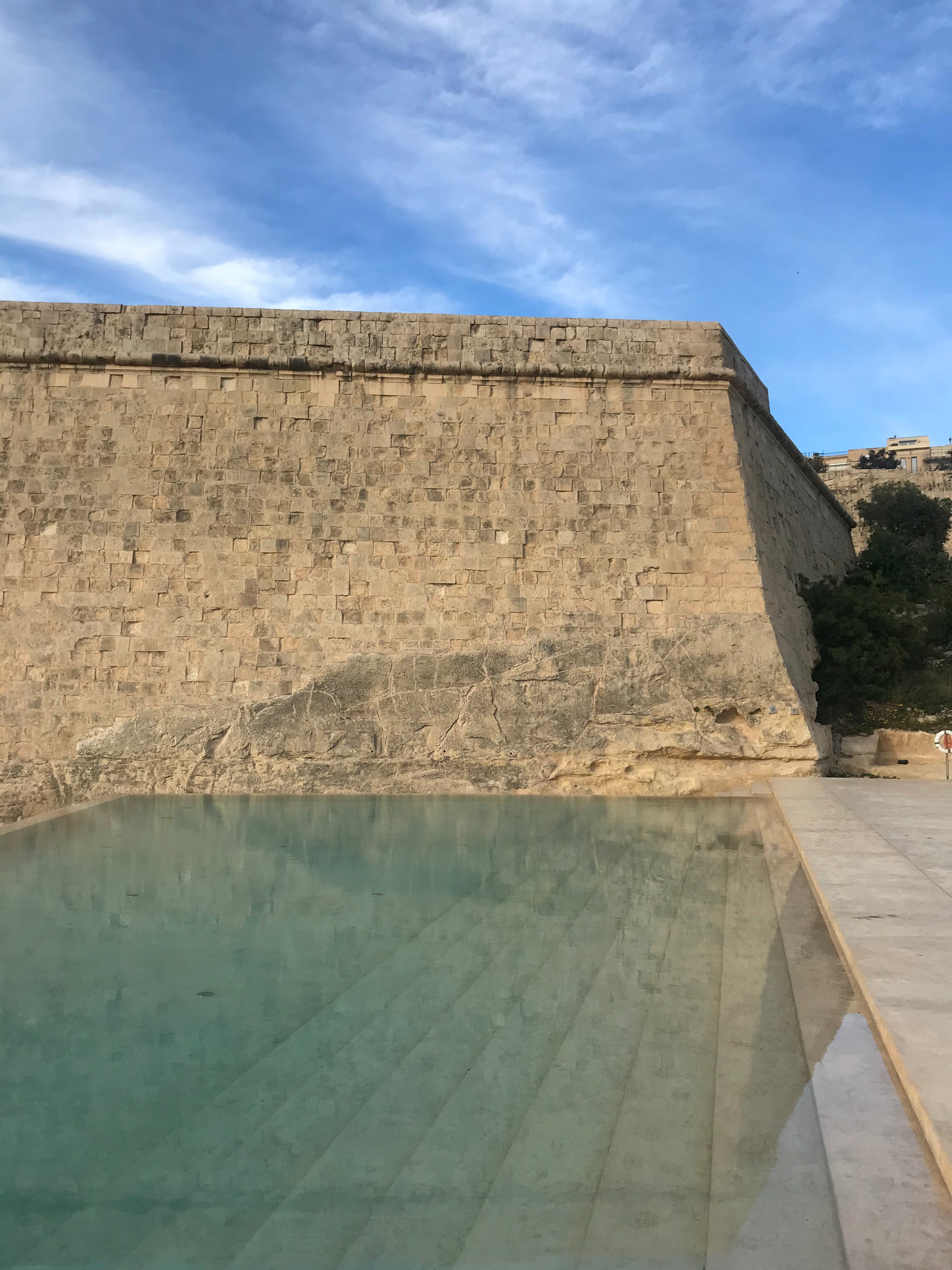 A large stone wall rises above a shallow reflecting pool with clear water, set under a blue sky with wispy clouds. Steps lead into the pool, and greenery is visible along the right edge.