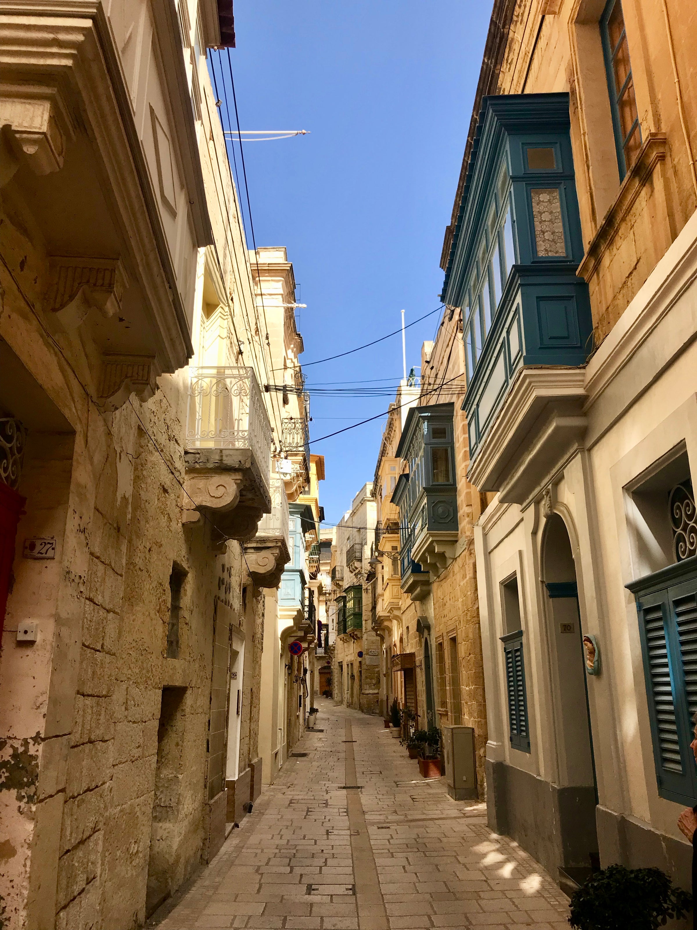 A narrow, sunlit street in a Mediterranean town, lined with yellow stone buildings featuring colorful balconies and shutters. The sky is clear and blue, and the street is empty.