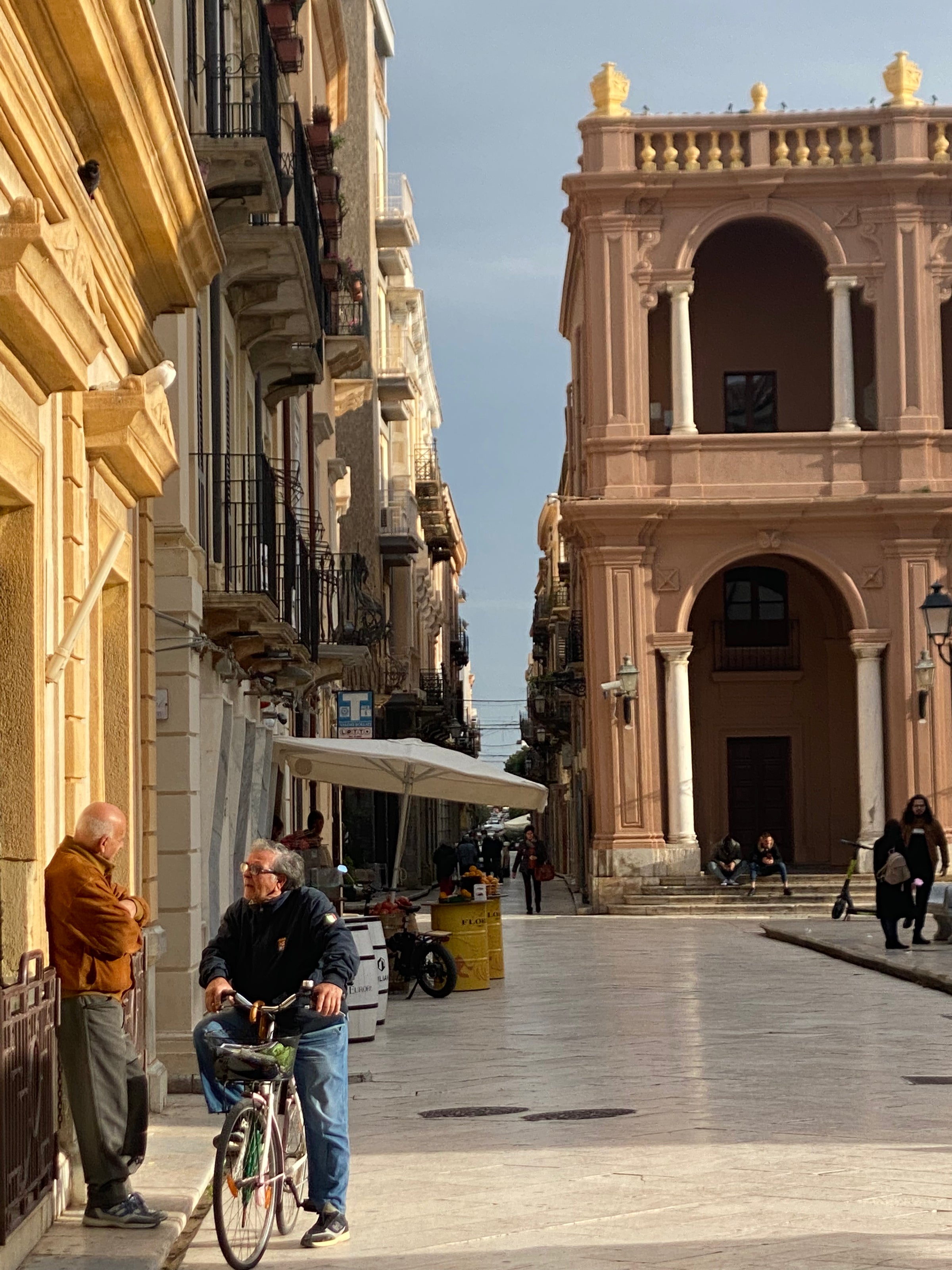 A man on a bicycle talks to another man standing on a narrow, sunlit street lined with old buildings and a grand arched structure in the background. People walk further down the street.