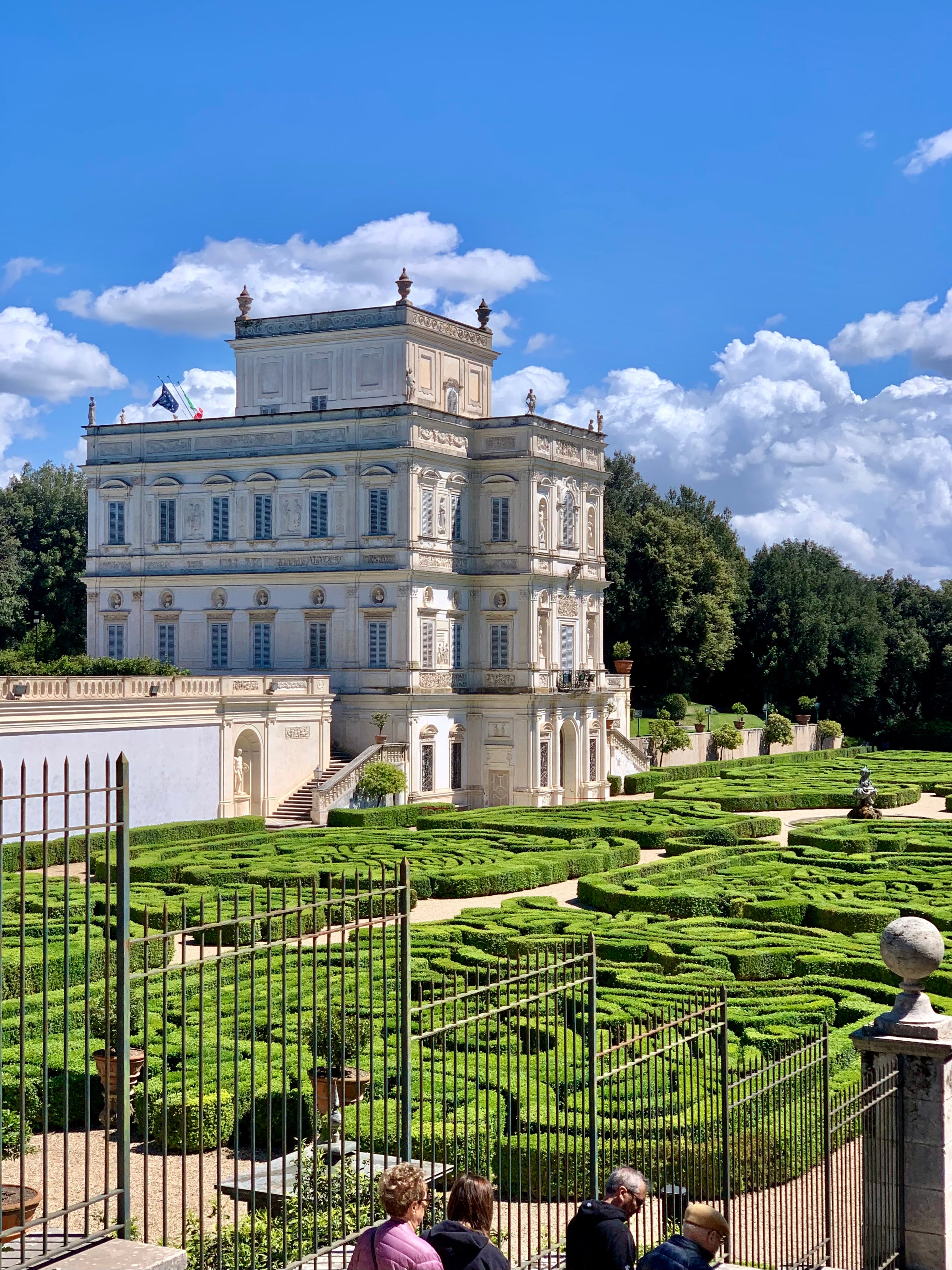 A grand, white three-story villa stands beside manicured hedge gardens under a bright blue sky with puffy clouds. Several people observe the scene from behind an iron fence in the foreground.