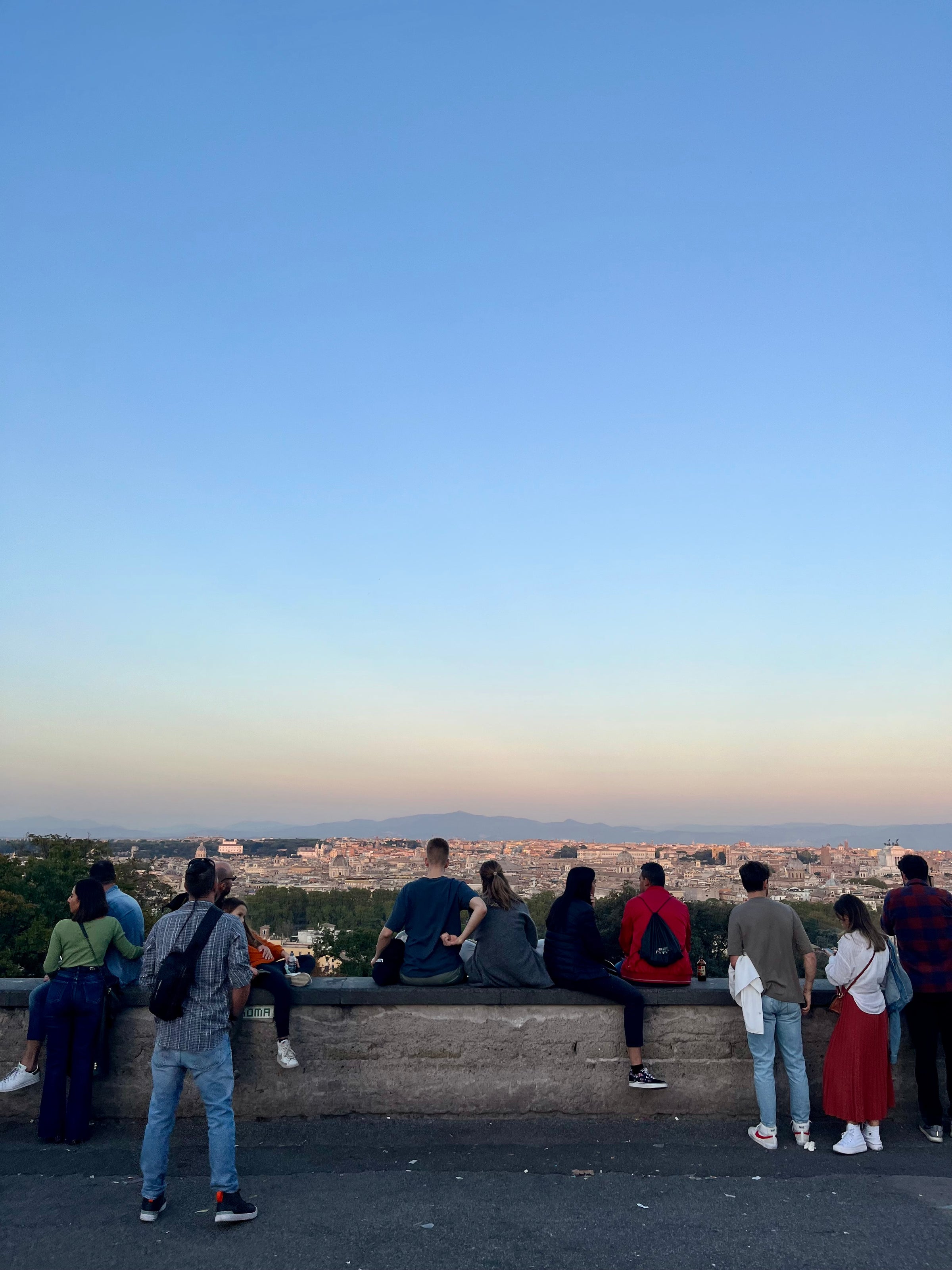 A group of people stand or sit on a stone wall, looking at a cityscape under a clear blue sky at sunset. The city stretches out in the distance, bathed in warm, golden light.