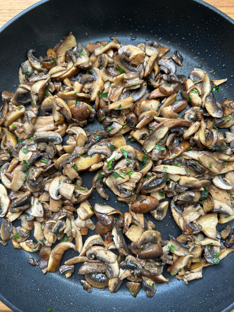 Sliced mushrooms sautéed with herbs and seasonings in a black frying pan, showing a mix of brown and white mushroom pieces on a wooden surface.