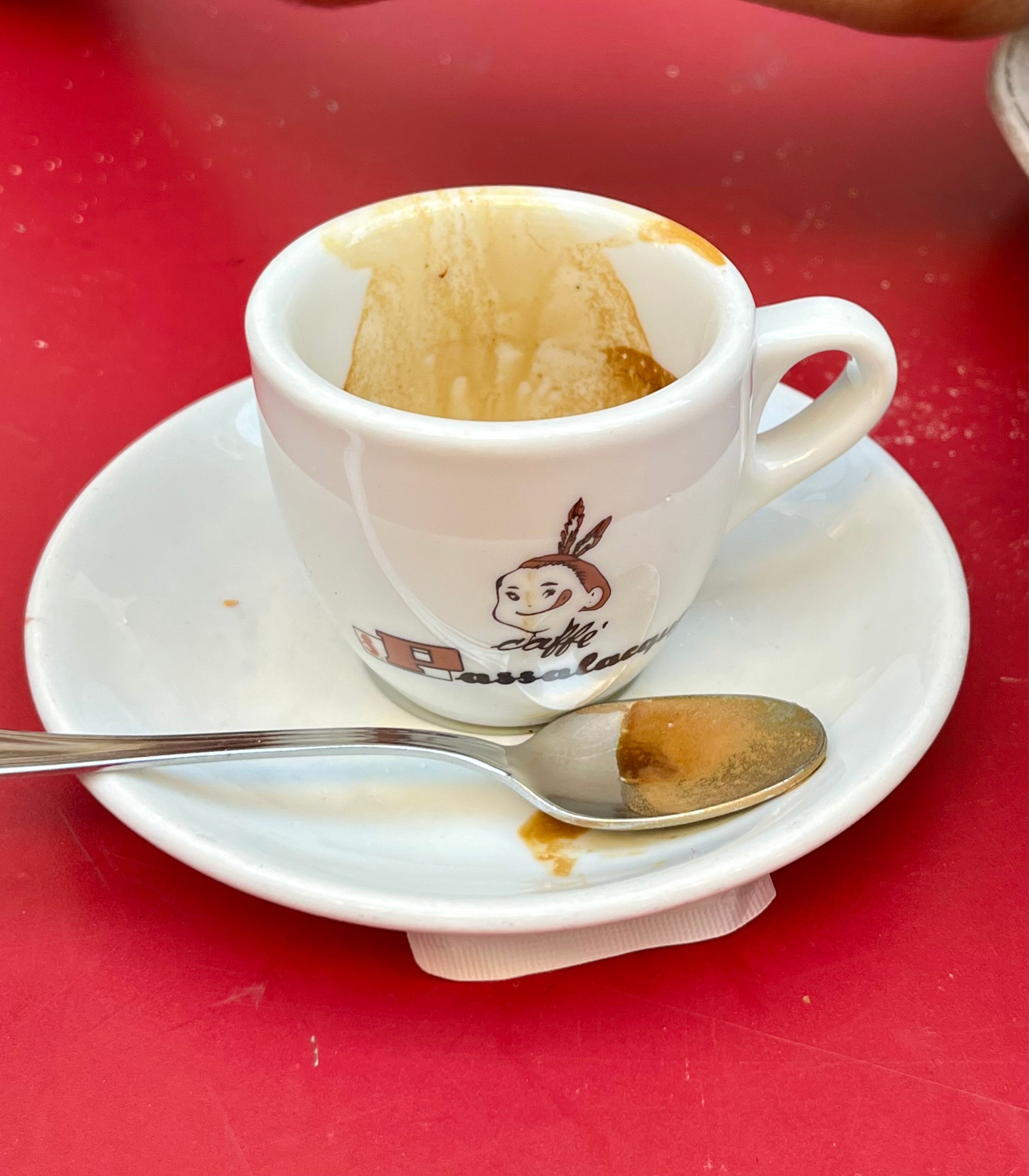 An empty espresso cup with coffee stains sits on a white saucer next to a spoon with coffee residue, against a red table background. The cup has a cartoon logo of a rabbit or animal wearing a headband.