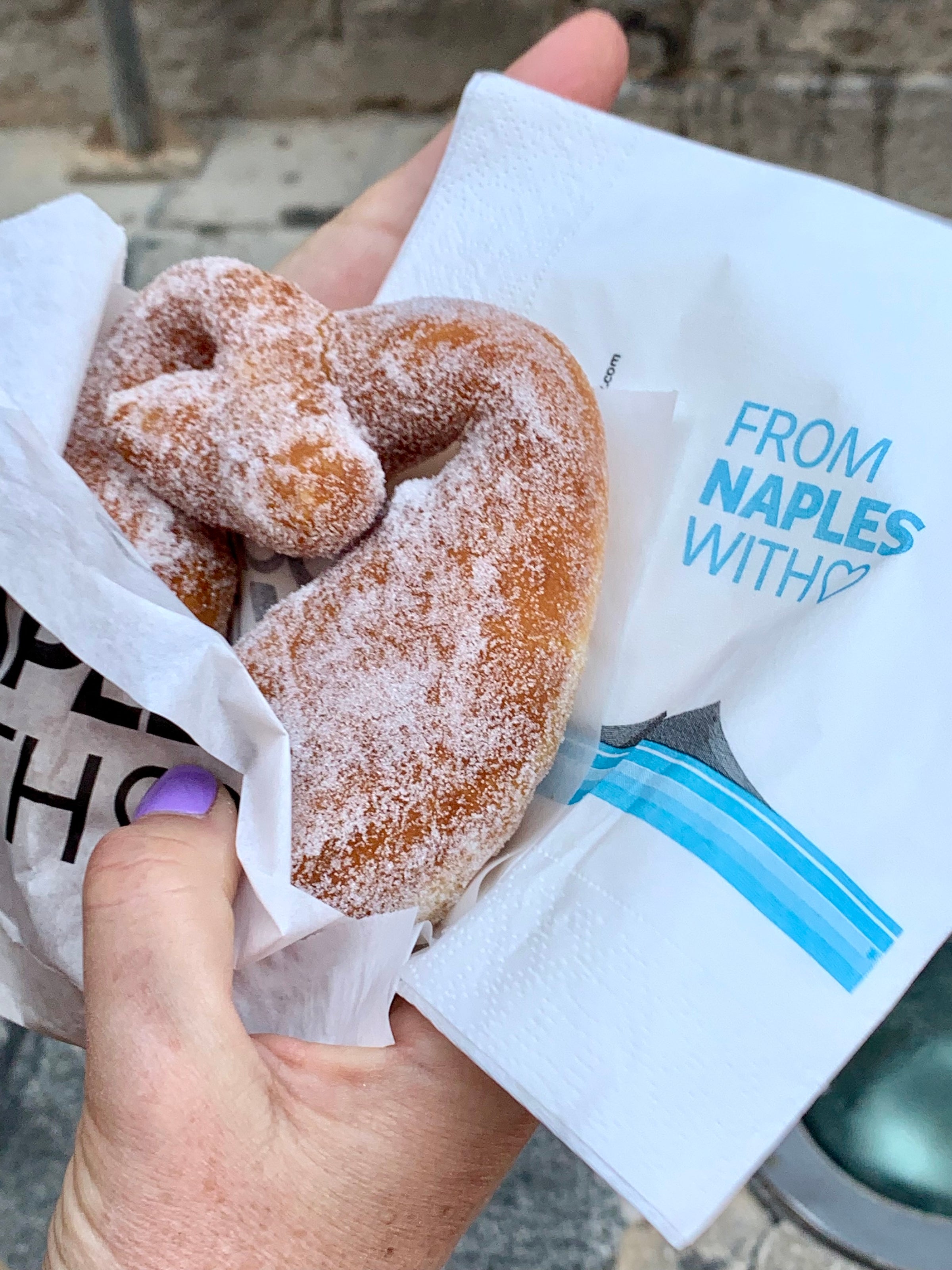 A hand holding a sugar-coated, pretzel-shaped pastry wrapped in paper, next to a white napkin with blue text that reads FROM NAPLES WITH ♥.