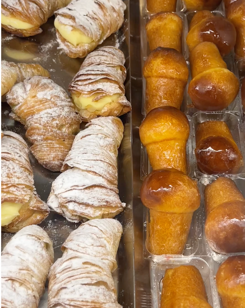 A tray of custard-filled sfogliatelle pastries dusted with powdered sugar is next to a tray of shiny, golden-brown baba au rhum cakes in a bakery display.