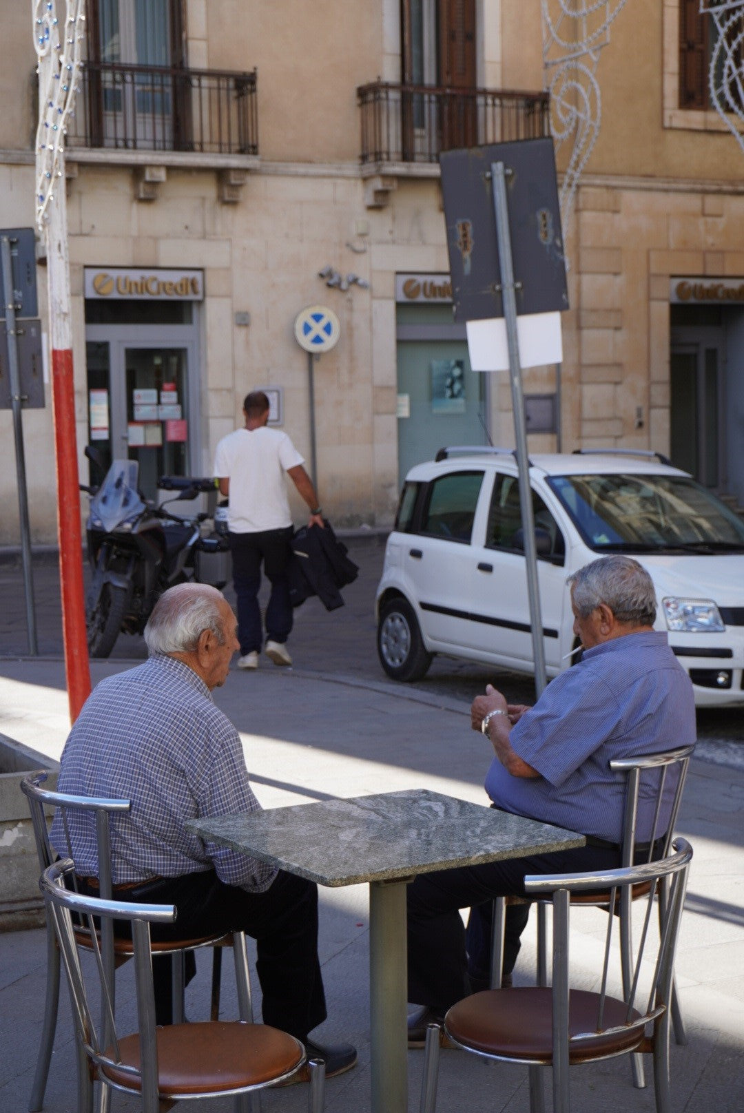 Two older men sit and talk at an outdoor table on a city street, while a man walks away in the background past parked cars and a bank. The scene is set in sunlight in front of a stone building.