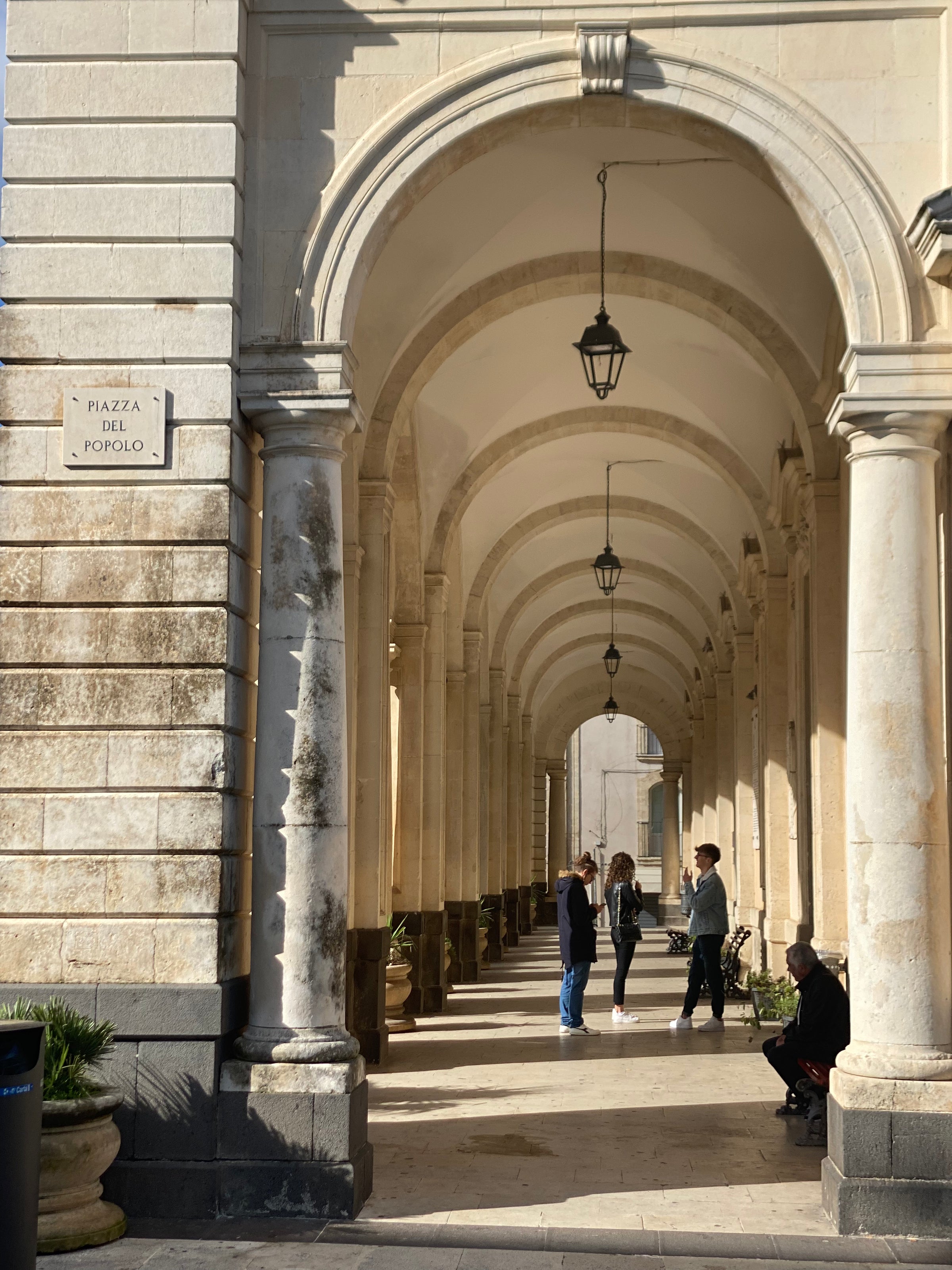 A sunlit stone archway with several arches in a row, hanging lanterns, and four people standing and talking under the arches. A sign on the wall reads Piazza del Popolo. One person sits near the entrance.