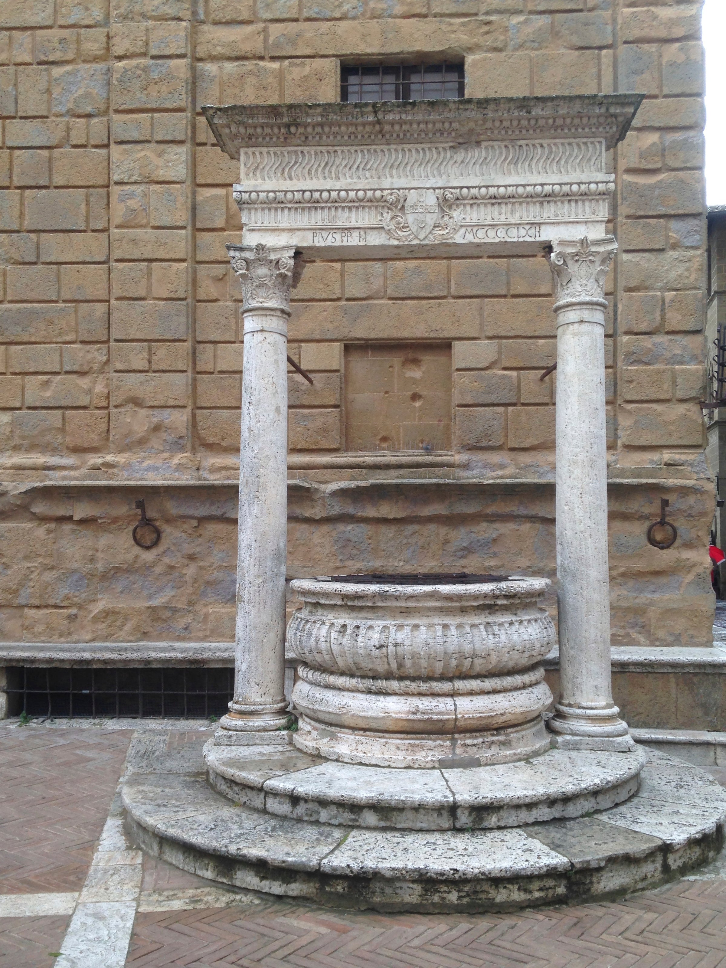 An old stone well with ornate carvings and two columns supporting a decorative top stands in a paved courtyard against a large brick wall.