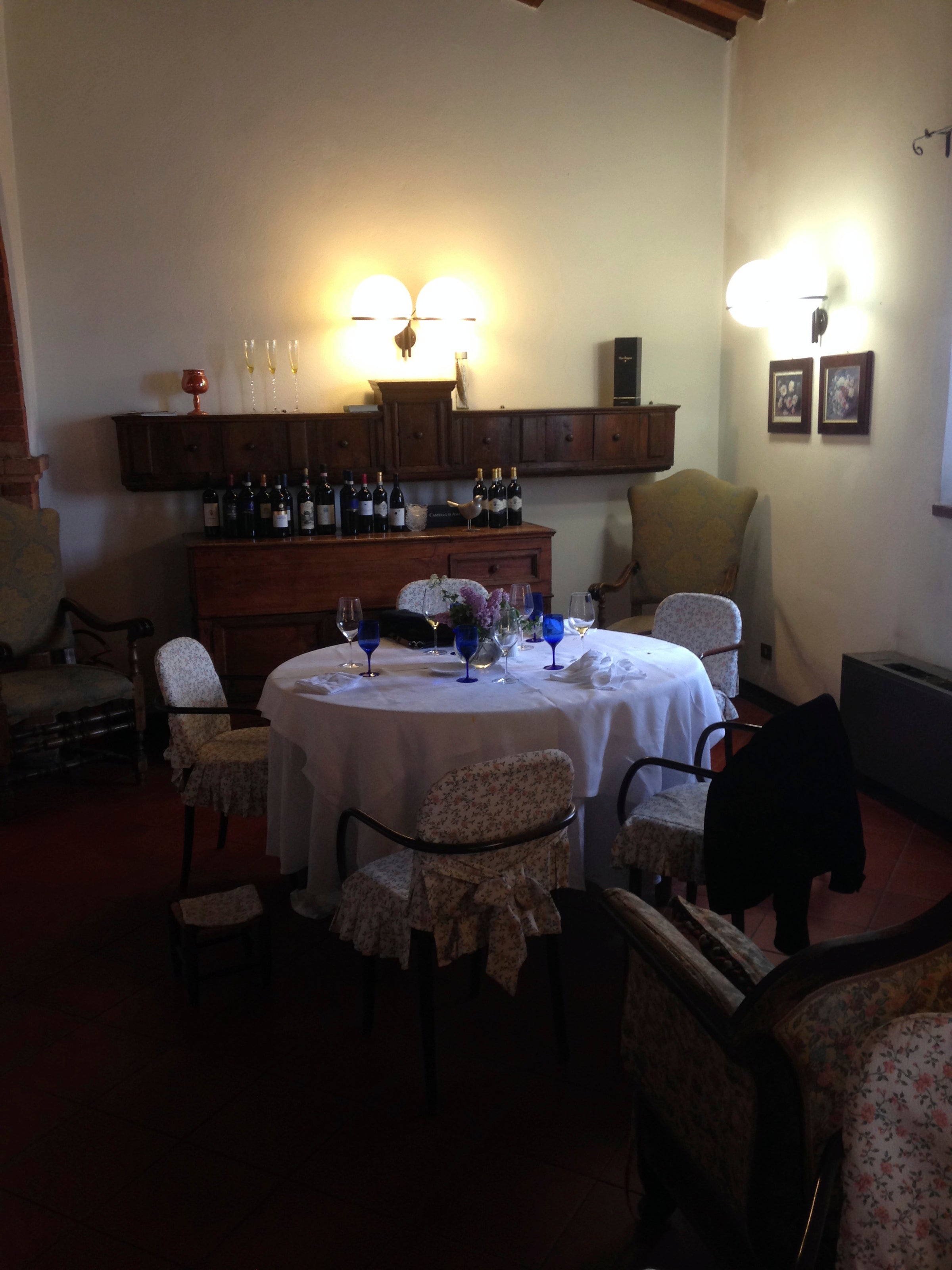 A round dining table with a white tablecloth is set for four in a cozy, dimly lit room; wooden chairs with floral covers surround the table, and shelves with wine bottles and glasses are in the background.