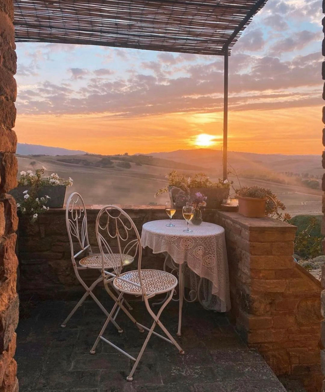 A cozy terrace with two ornate white chairs and a small round table set for two, overlooking rolling hills at sunset. Two glasses of wine sit on the table, with flowers and potted plants decorating the rustic space.
