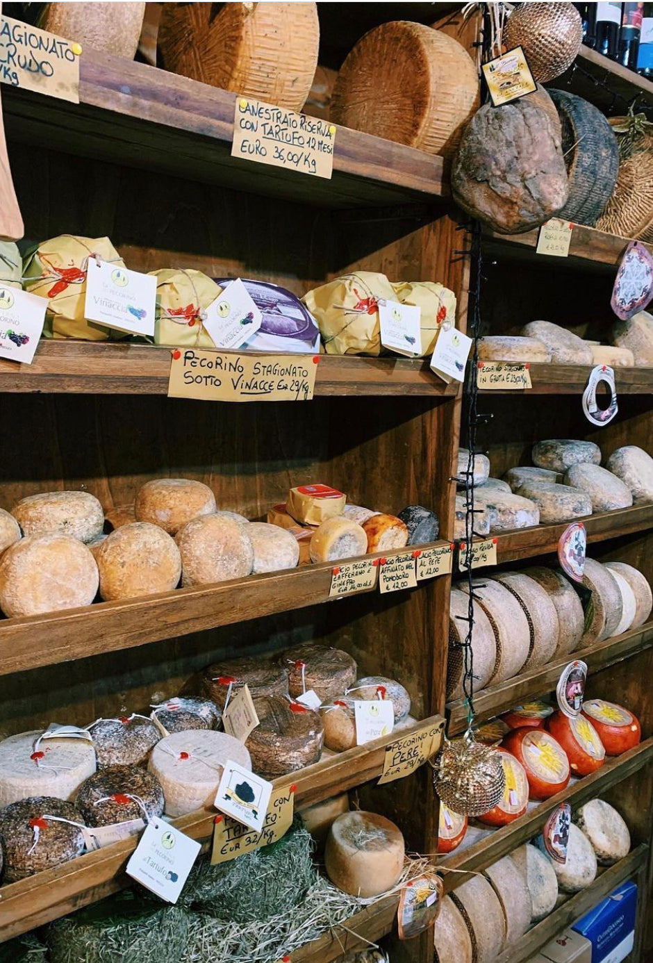Shelves filled with assorted Italian cheeses, some wrapped in paper or wax, others uncovered. Various handwritten labels with prices and names are attached, creating a rustic, artisanal market display.