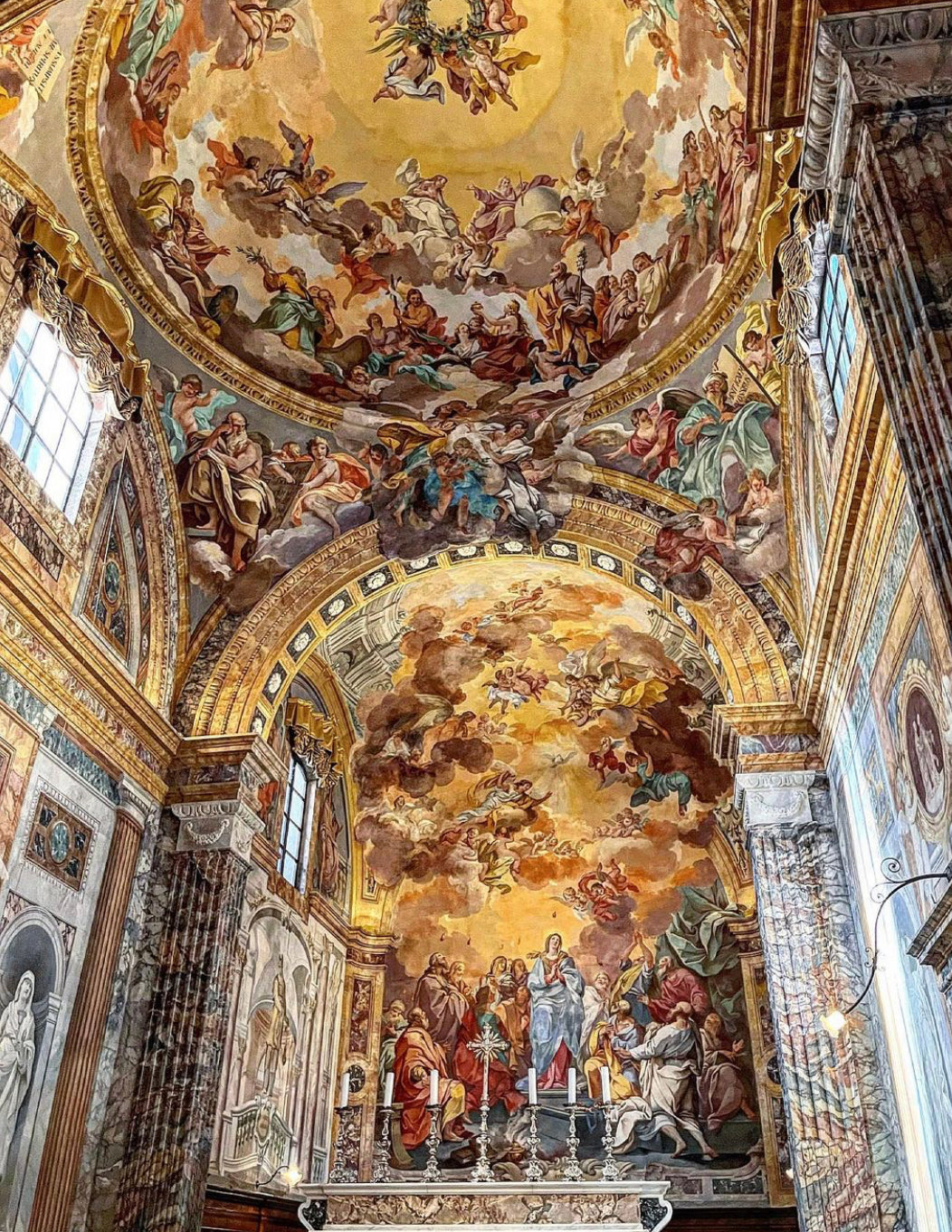 Elaborate frescoes cover the arched ceiling and upper walls of a church, depicting religious scenes with numerous figures, vibrant colors, and intricate architectural details under natural light from tall windows.