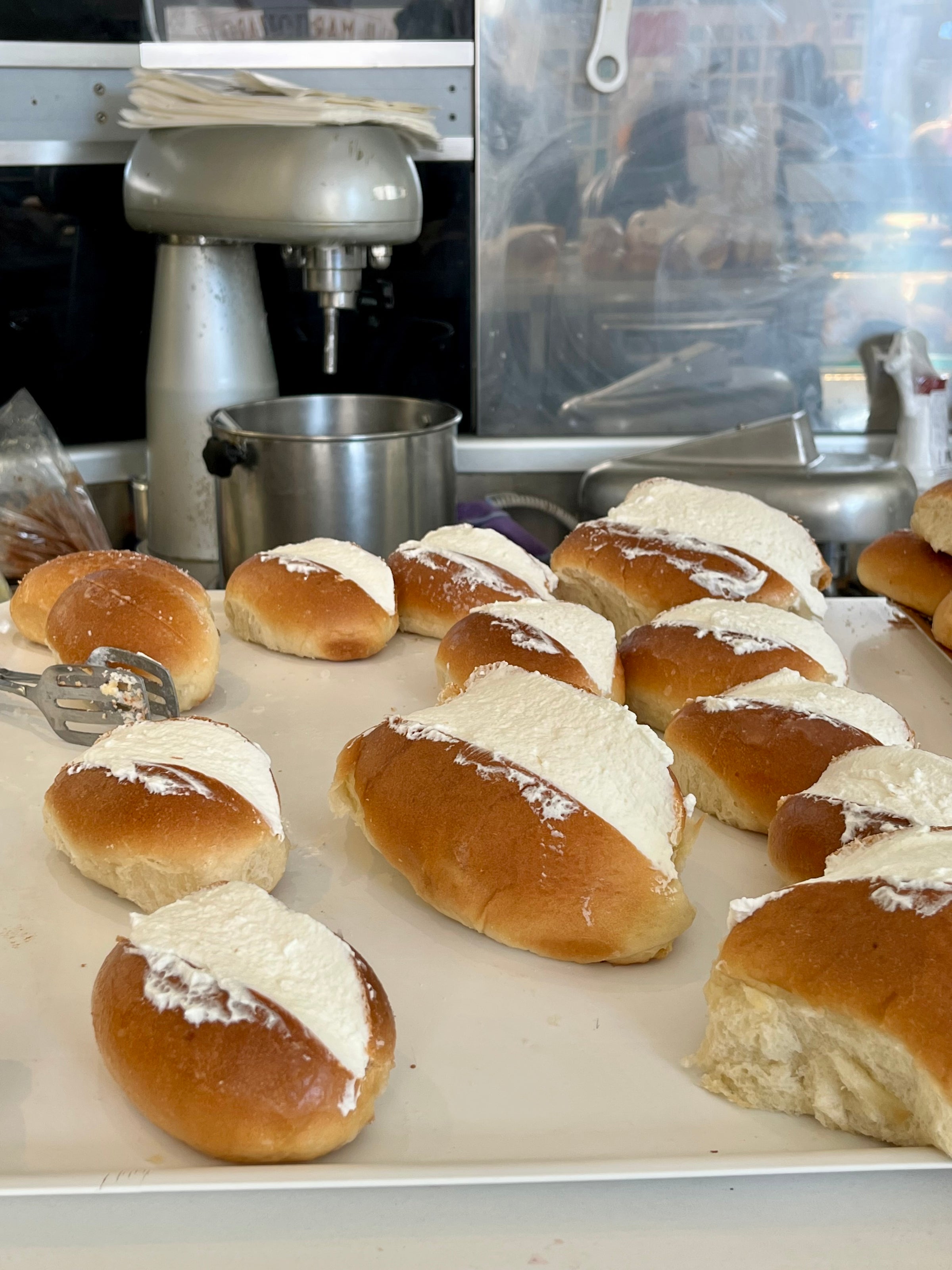 A tray of freshly baked brioche buns filled with whipped cream, displayed on a counter in a bakery kitchen with baking equipment in the background.