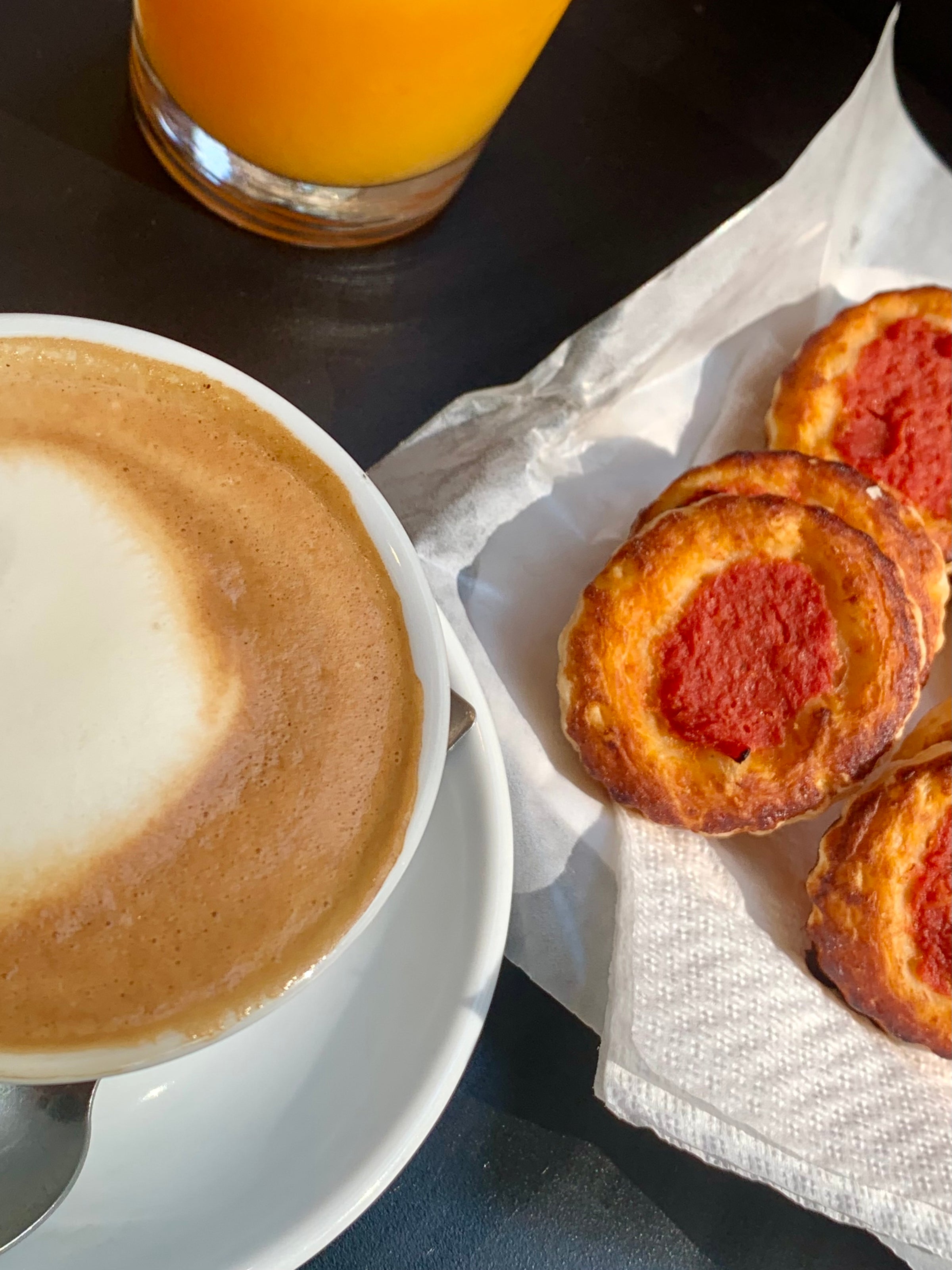 A cup of cappuccino on a saucer with a spoon, next to three small pastries topped with tomato sauce on a napkin, and a glass of orange juice in the background.