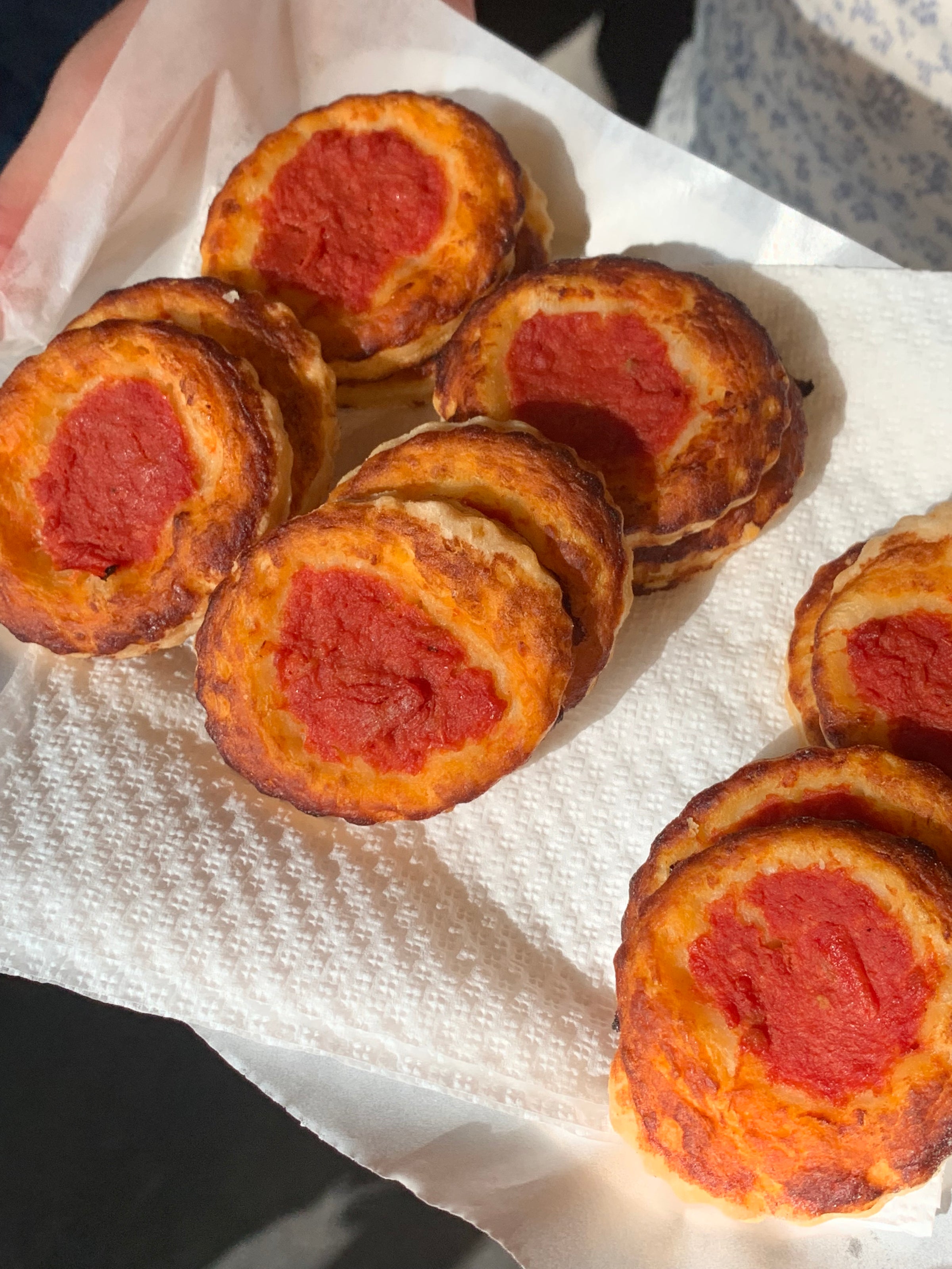 Six small, round savory pastries with golden-brown edges and tomato paste centers are arranged on a white paper towel, with sunlight shining on them.