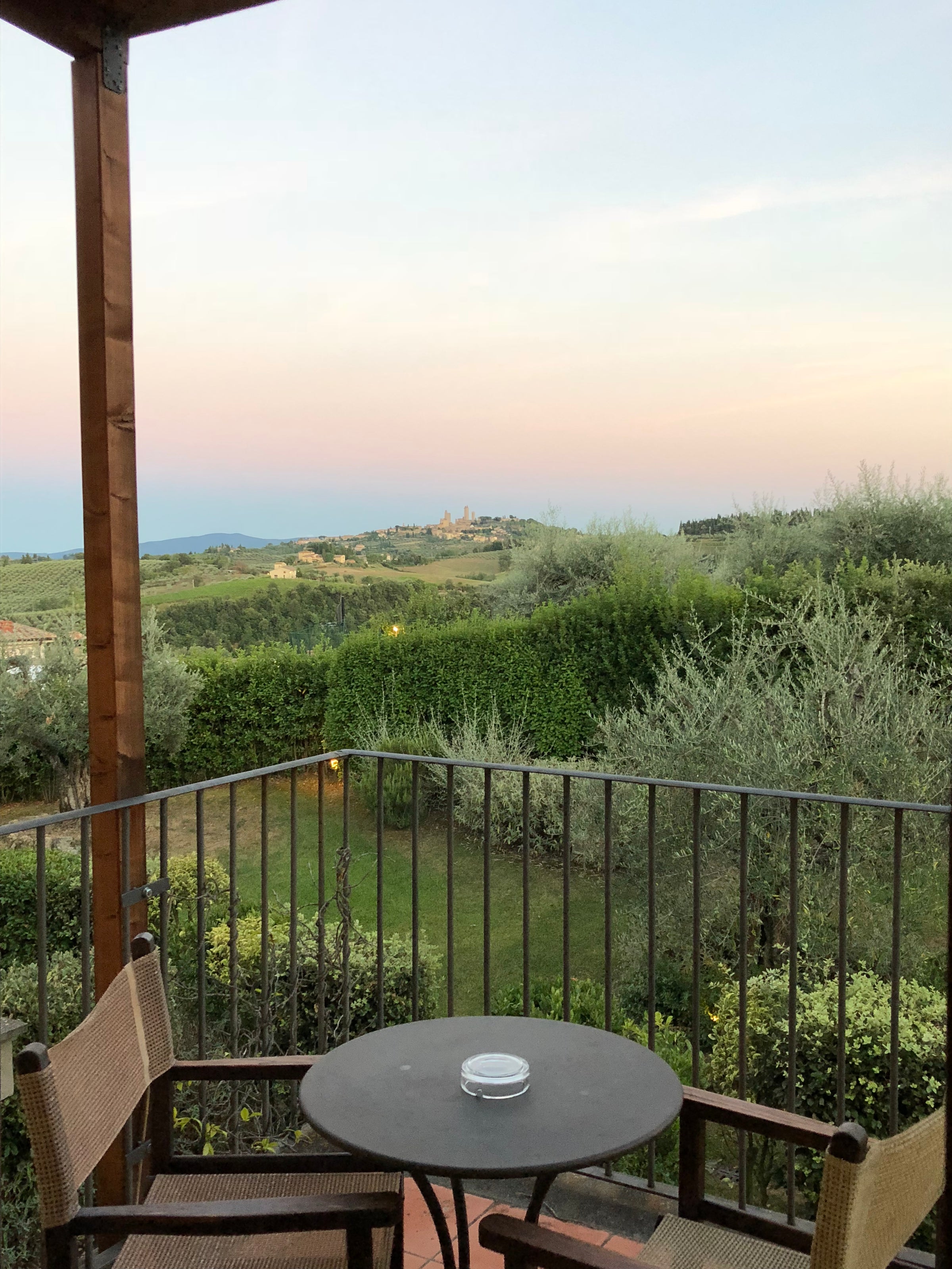 A small round table with two chairs on a patio overlooks lush green hedges, olive trees, and a distant village under a pastel sunset sky. A railing surrounds the patio.