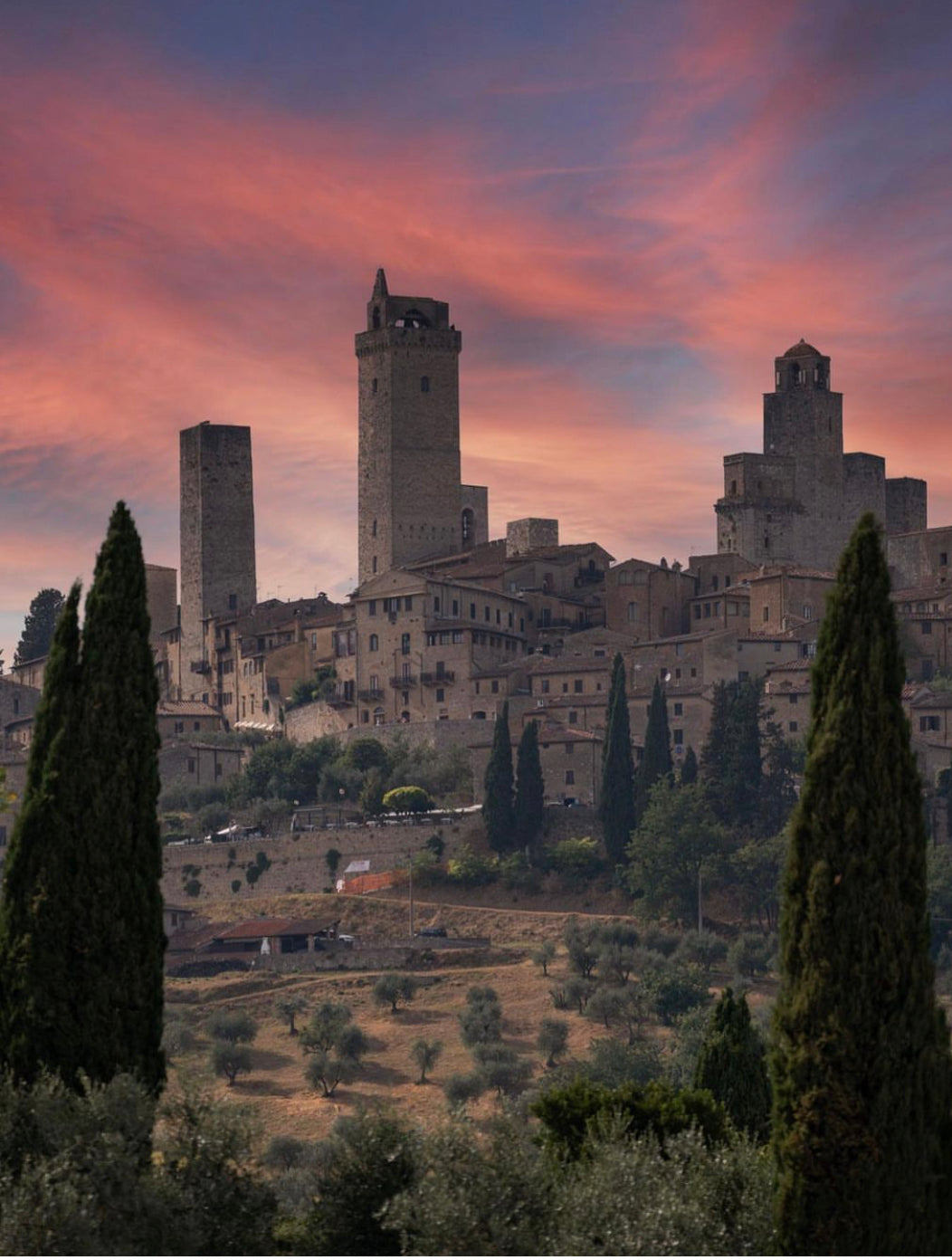 Medieval stone towers rise above the hillside town of San Gimignano, Italy, with tall cypress trees in the foreground and a dramatic pink and purple sunset sky in the background.