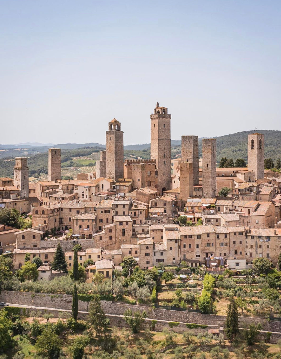 A medieval Italian town with numerous tall stone towers rises above tan buildings, surrounded by green hills and trees under a clear sky.