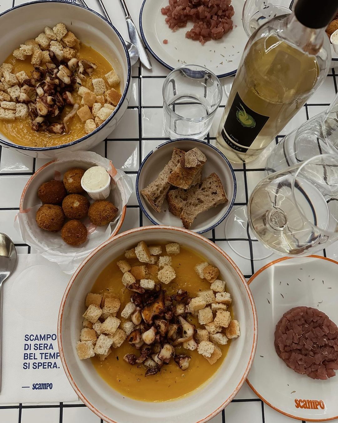 A table set for a meal with bowls of creamy soup topped with croutons and octopus, plates of bread and falafel with sauce, a bottle of white wine, filled glasses, and a card with Italian text.