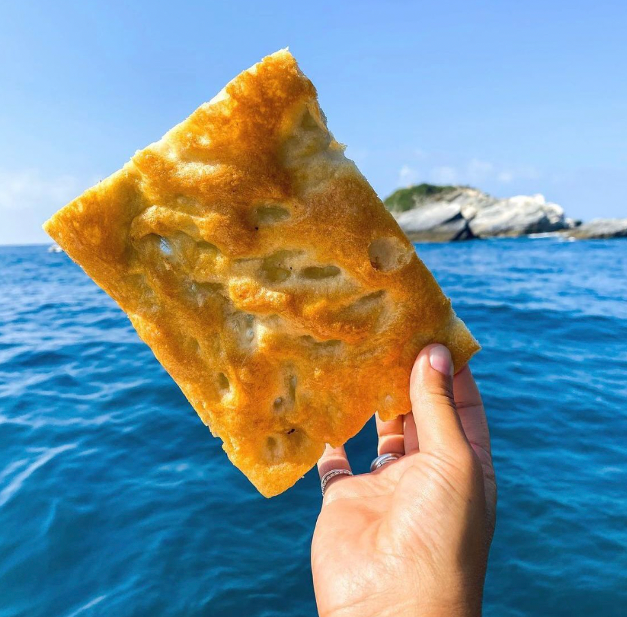 A hand holds up a rectangular piece of golden, airy focaccia bread against a backdrop of blue ocean and a distant rocky island under a clear sky.