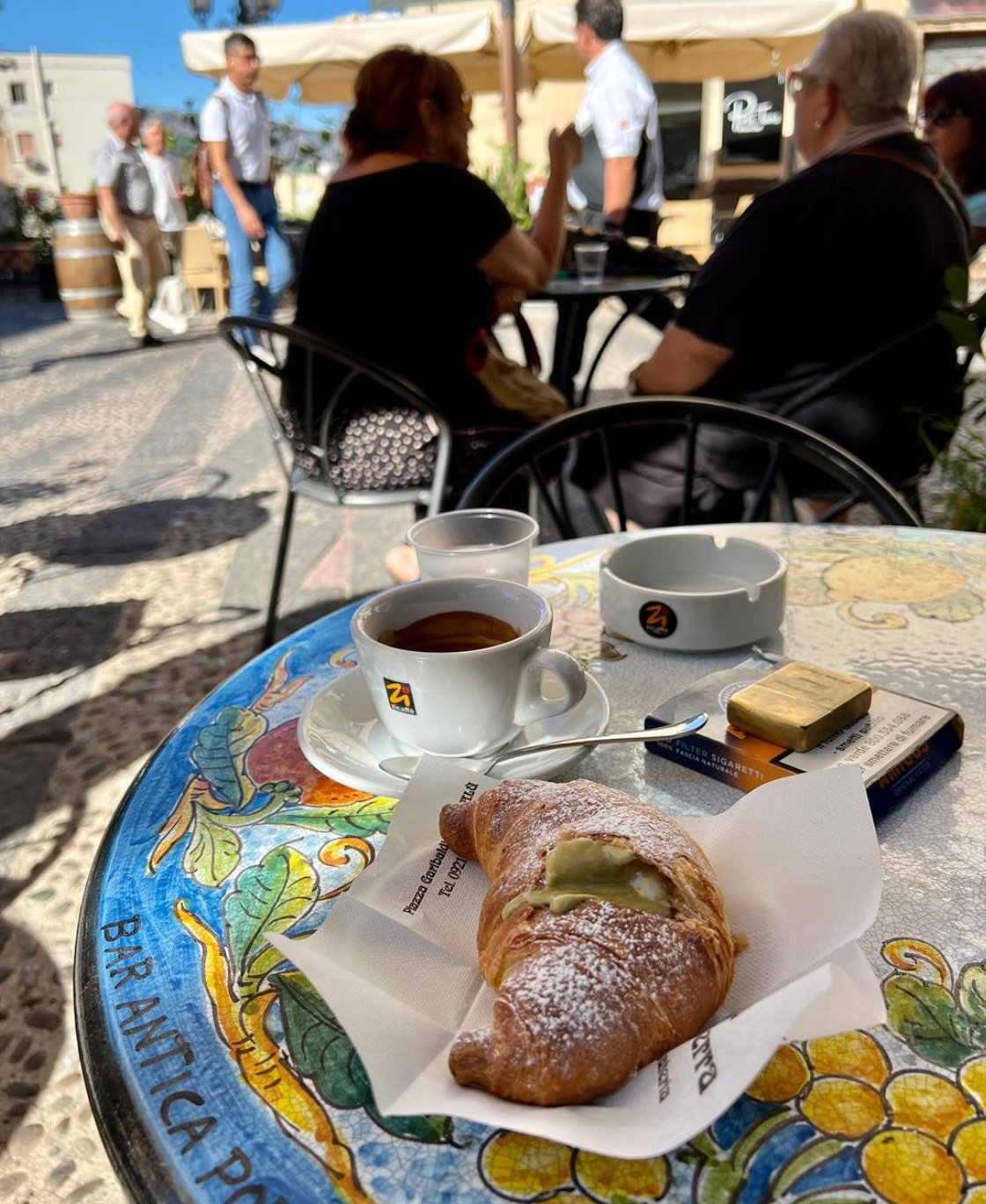 A table at an outdoor café with a cappuccino, an ashtray, a lighter, and a pastry dusted with powdered sugar. People are sitting and walking in the sunlit background on a mosaic-tiled patio.