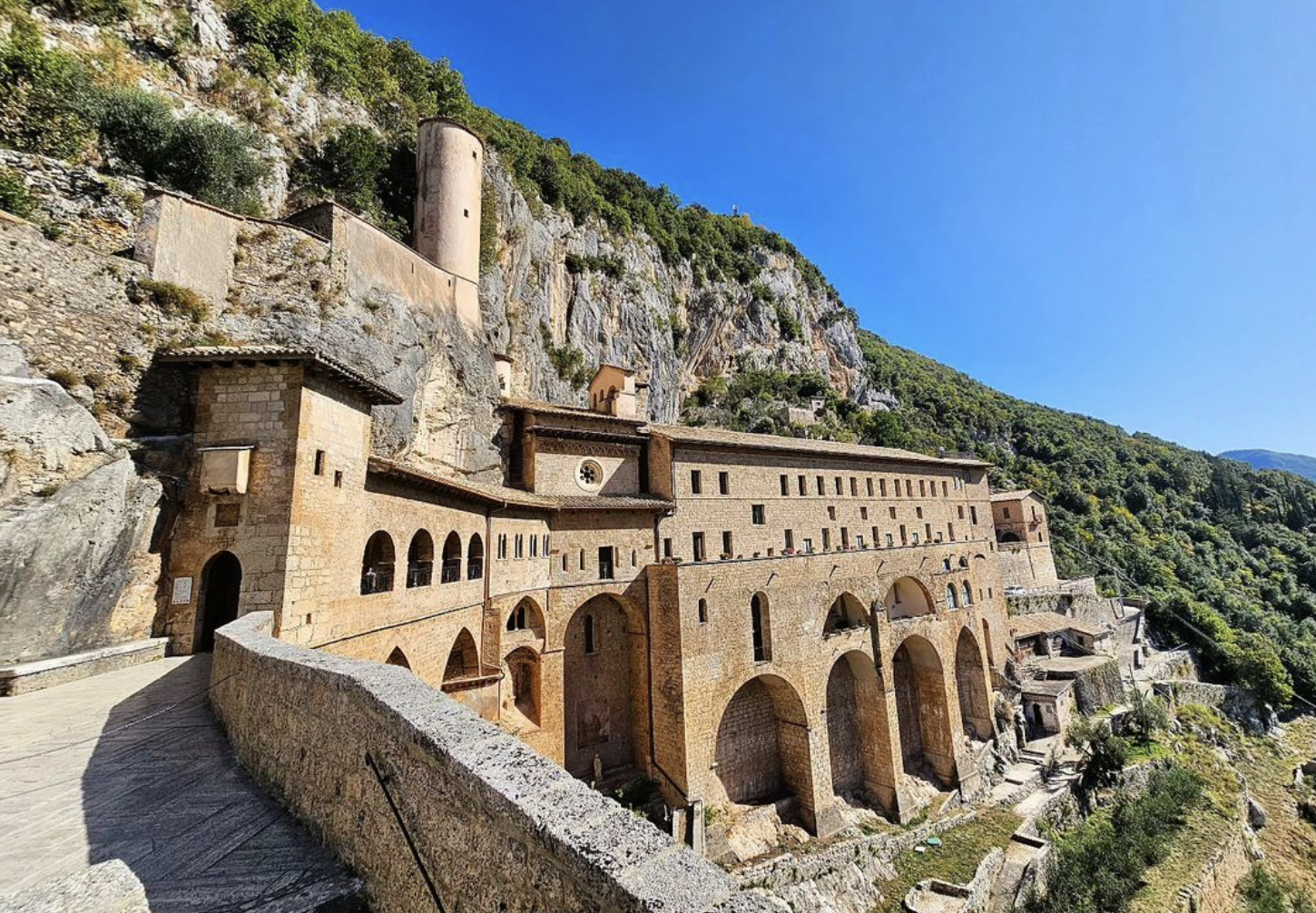 A historic stone monastery with arches and a tower is built into a rocky hillside, surrounded by lush green trees under a clear blue sky.