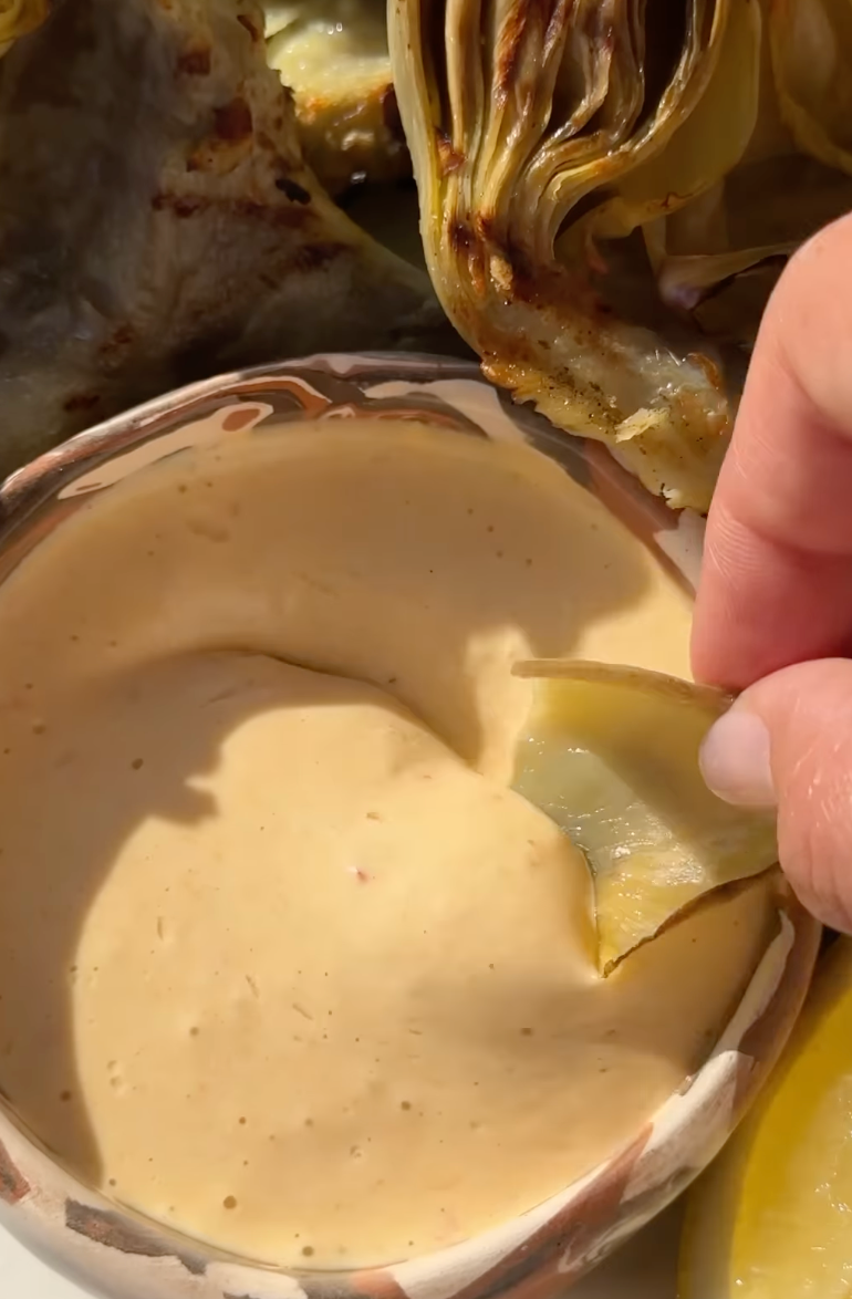 A hand dips an artichoke leaf into a creamy, light orange sauce in a small bowl, with more artichoke pieces visible in the background.