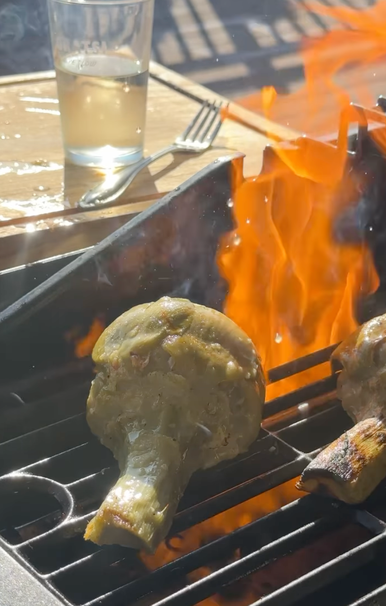 A large artichoke is grilling over open flames on a barbecue. In the background, there is a glass of water, a fork, and sunlight reflecting on a wooden table.