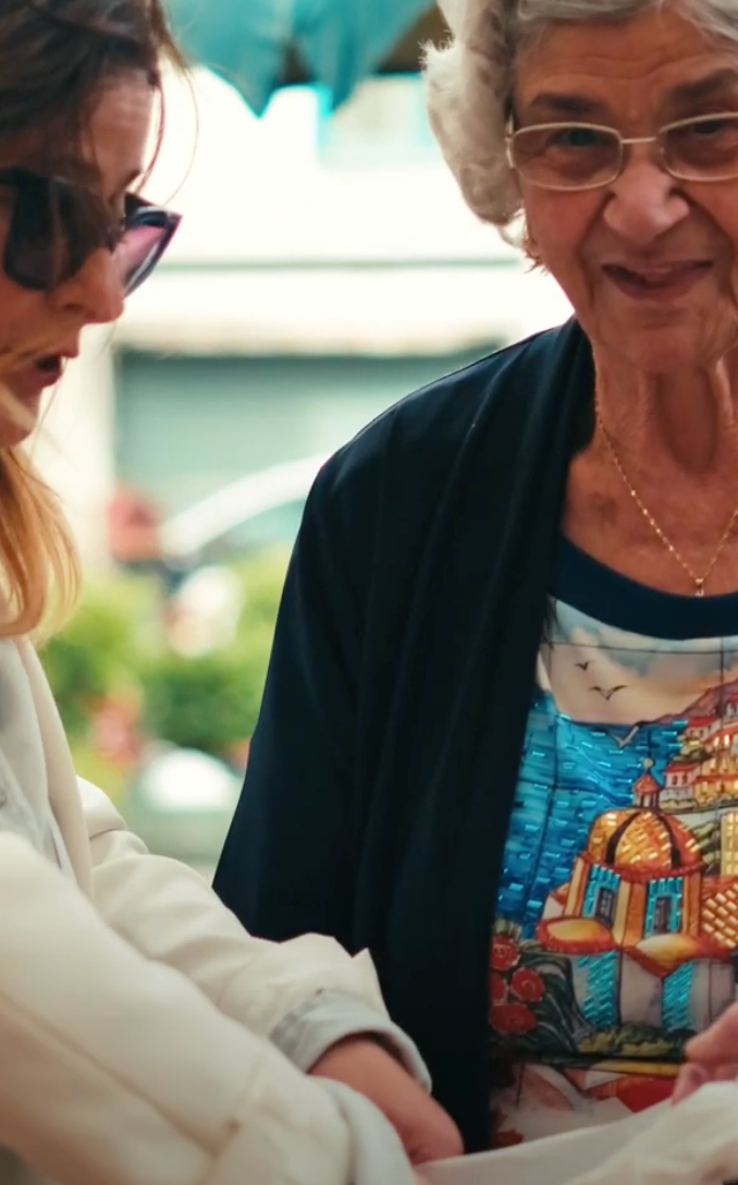 Two women standing together outdoors, one older and smiling, the other younger and wearing sunglasses. The older woman is wearing a colorful shirt with a scenic design. They appear to be engaged in conversation or an activity.