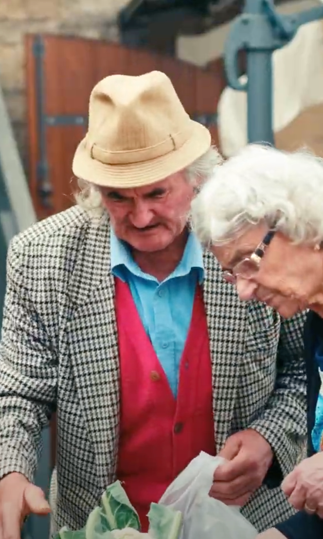 An elderly man in a beige hat and checkered jacket stands beside an elderly woman with white hair and glasses as they shop for produce at an outdoor market.