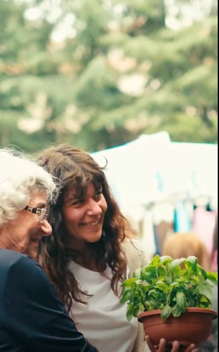 Two women, one older and one younger, smile together outdoors. The younger woman holds a small potted basil plant. Blurred greenery and tents are visible in the background, suggesting a market or outdoor event.