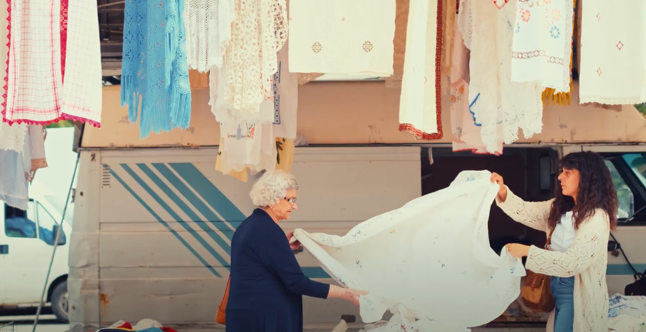 Two women hold up a large white cloth at an outdoor market, surrounded by hanging linens and fabric. A van with blue stripes is in the background. One woman has gray hair; the other has long dark hair.