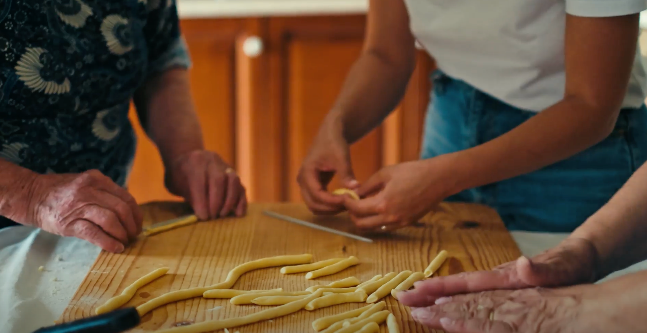 Three people shape fresh pasta dough into long pieces on a wooden board. Their faces are out of frame, focusing on their hands as they work together in a warm kitchen setting.