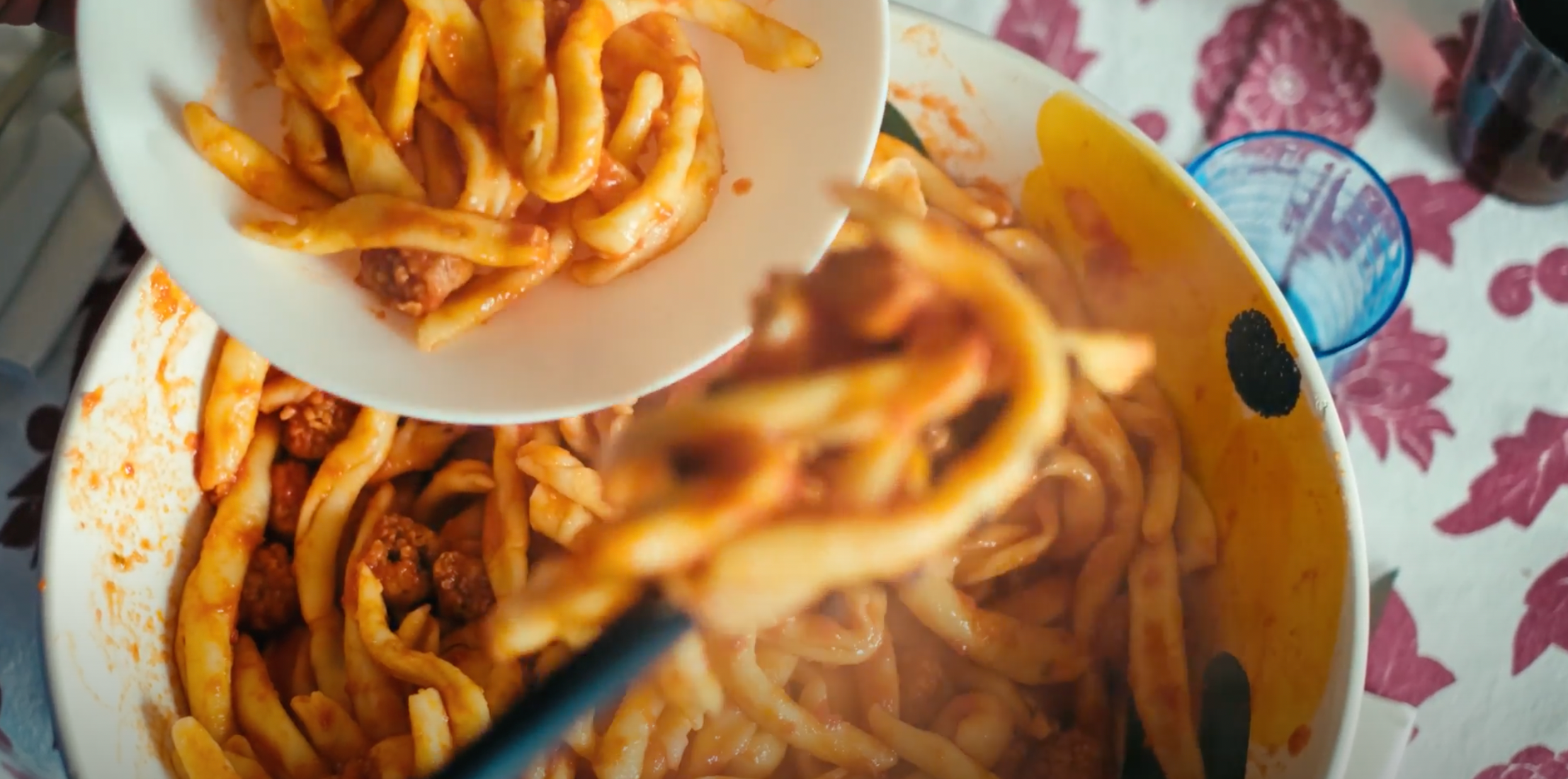 A serving of pasta with tomato sauce and meatballs is being spooned from a large pot into a white bowl on a patterned tablecloth.