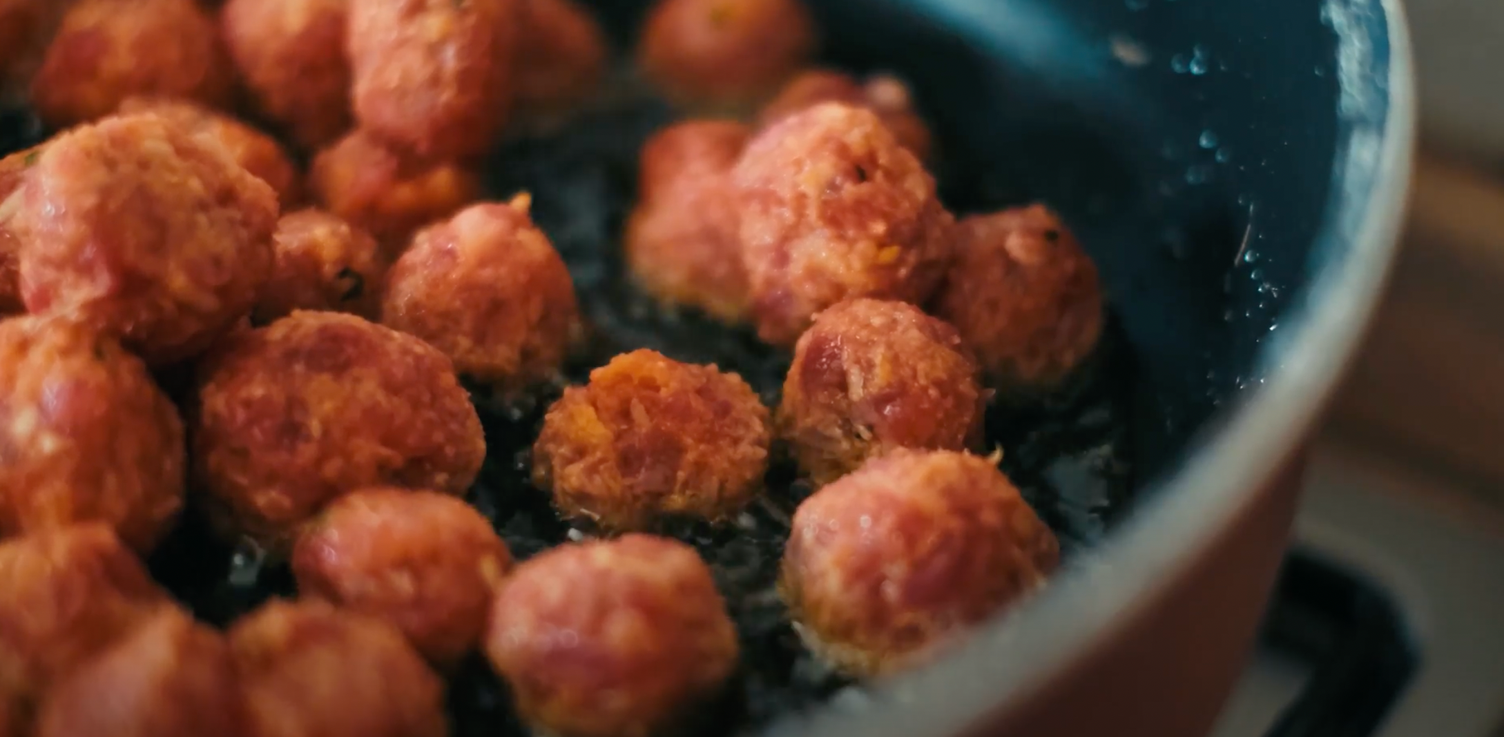 A close-up view of several meatballs frying in hot oil in a dark pan. The meatballs are golden brown and crispy, bubbles forming around them as they cook.