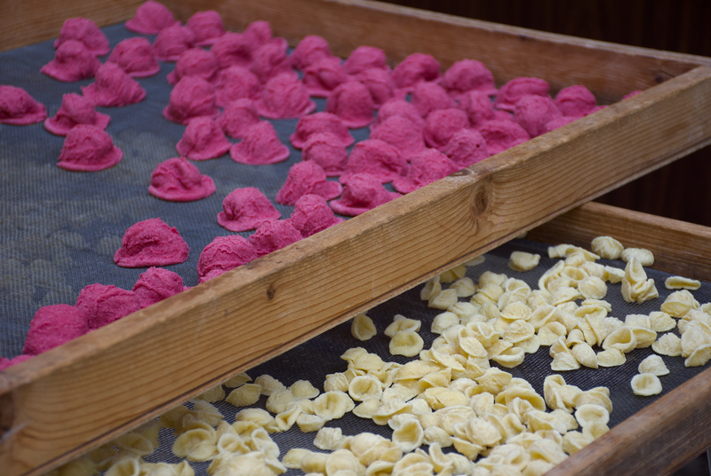 Two wooden trays with mesh bottoms are filled with uncooked pasta; the top tray holds pink pasta, while the bottom tray contains yellow pasta, both arranged in a single layer to dry.