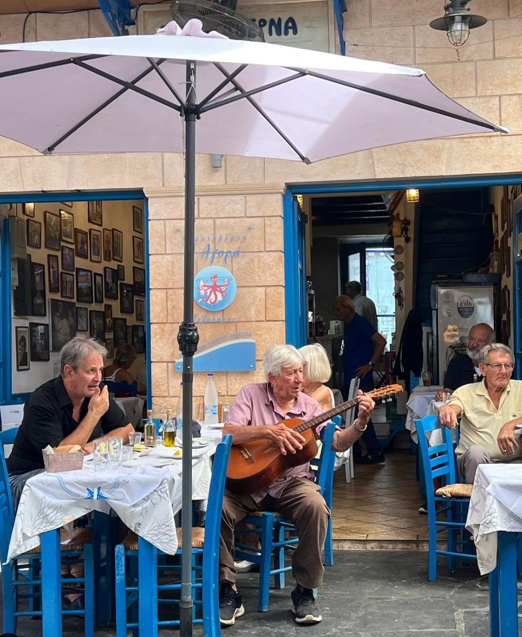 An older man plays a guitar and sings at an outdoor cafe with blue chairs and white tables, while others sit and listen under a large umbrella. The scene is lively and relaxed, set in front of a stone building.