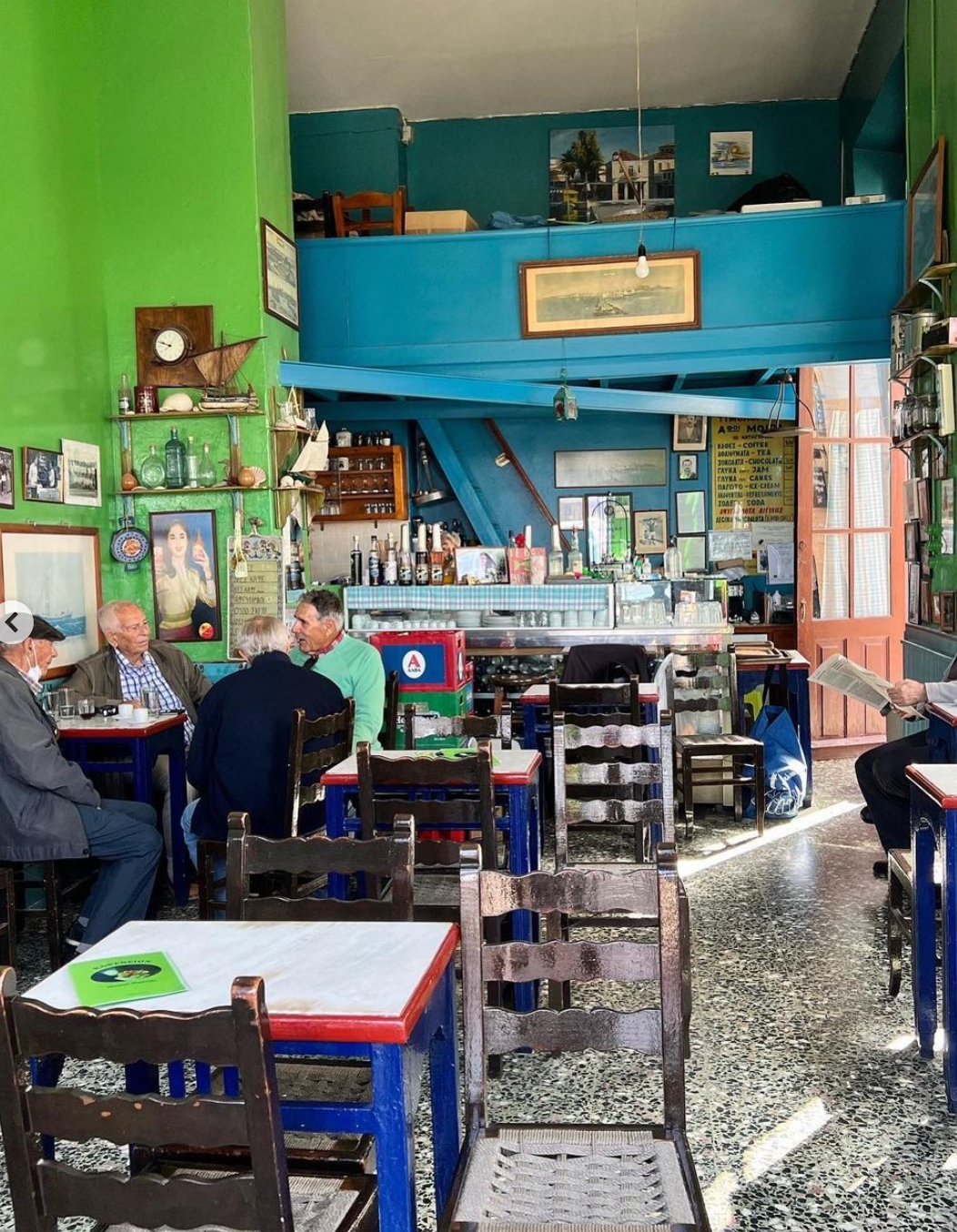 A colorful, sunlit café with green and blue walls, vintage decor, and framed photos. Several elderly men sit at tables chatting, while the bar and shelves of bottles are visible in the background.