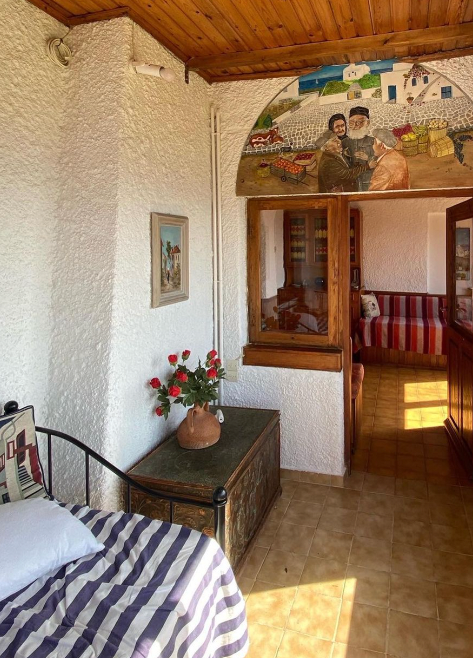 Cozy room with white textured walls, wooden ceiling, a striped bed, a chest with a vase of red and white flowers, wall art, and an archway featuring a mural of people and houses, leading to another room.