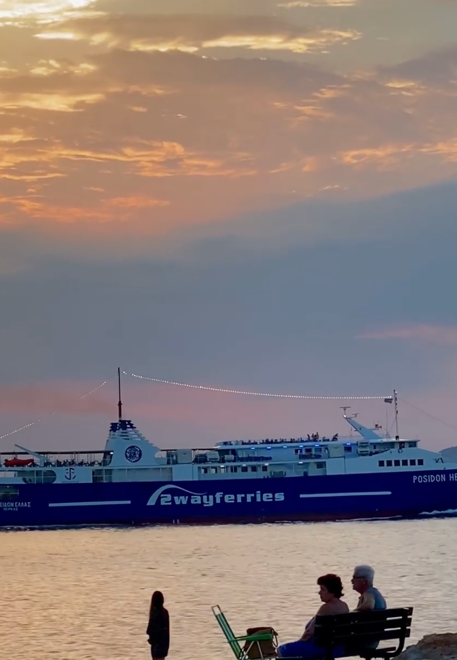 A large blue and white ferry labeled 2wayferries sails on calm water at sunset, with orange clouds in the sky and silhouettes of two people standing in the foreground.