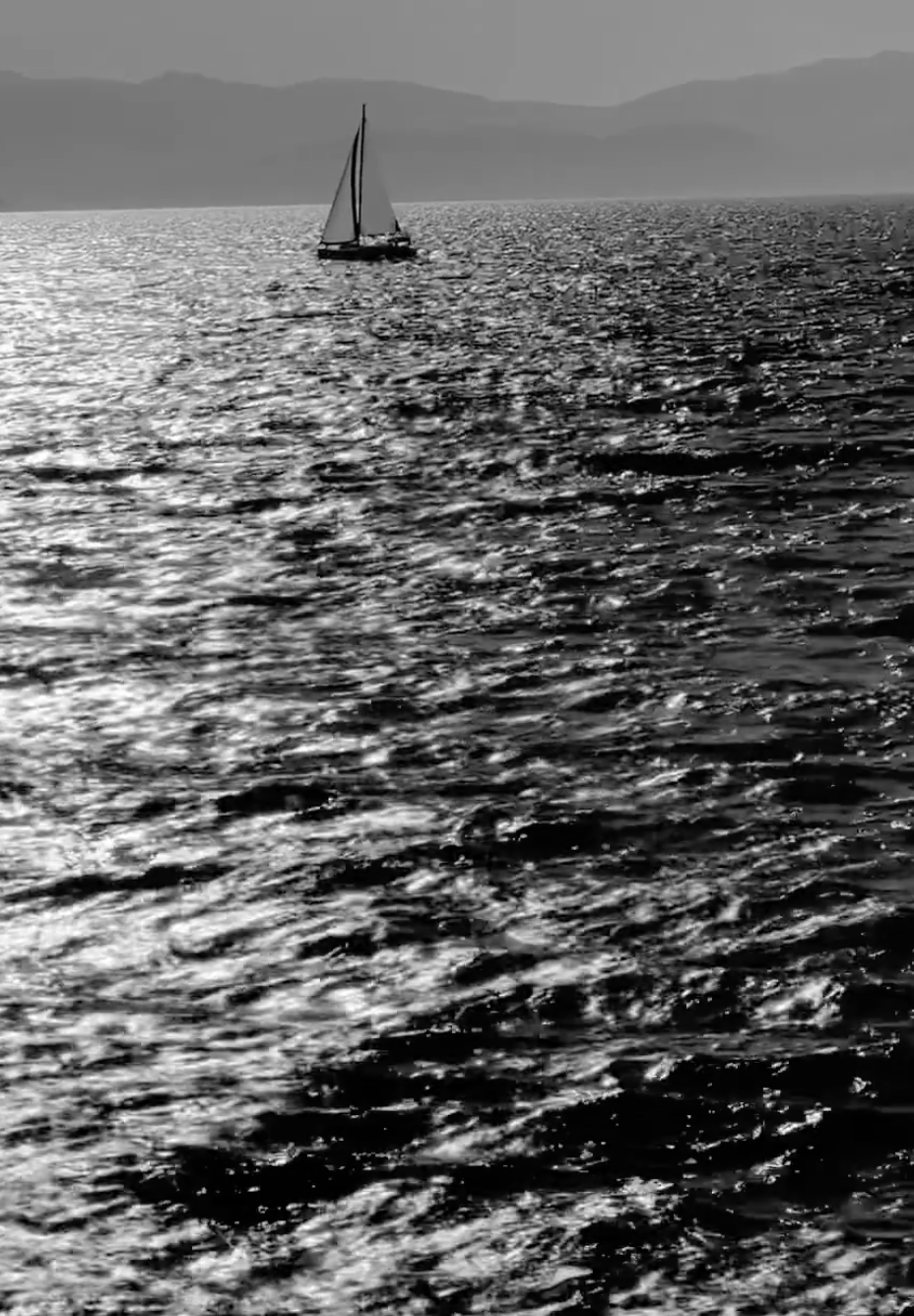 A sailboat glides across a shimmering, sunlit sea with gentle waves, while distant mountains form a hazy backdrop under a clear sky. The image is in black and white.