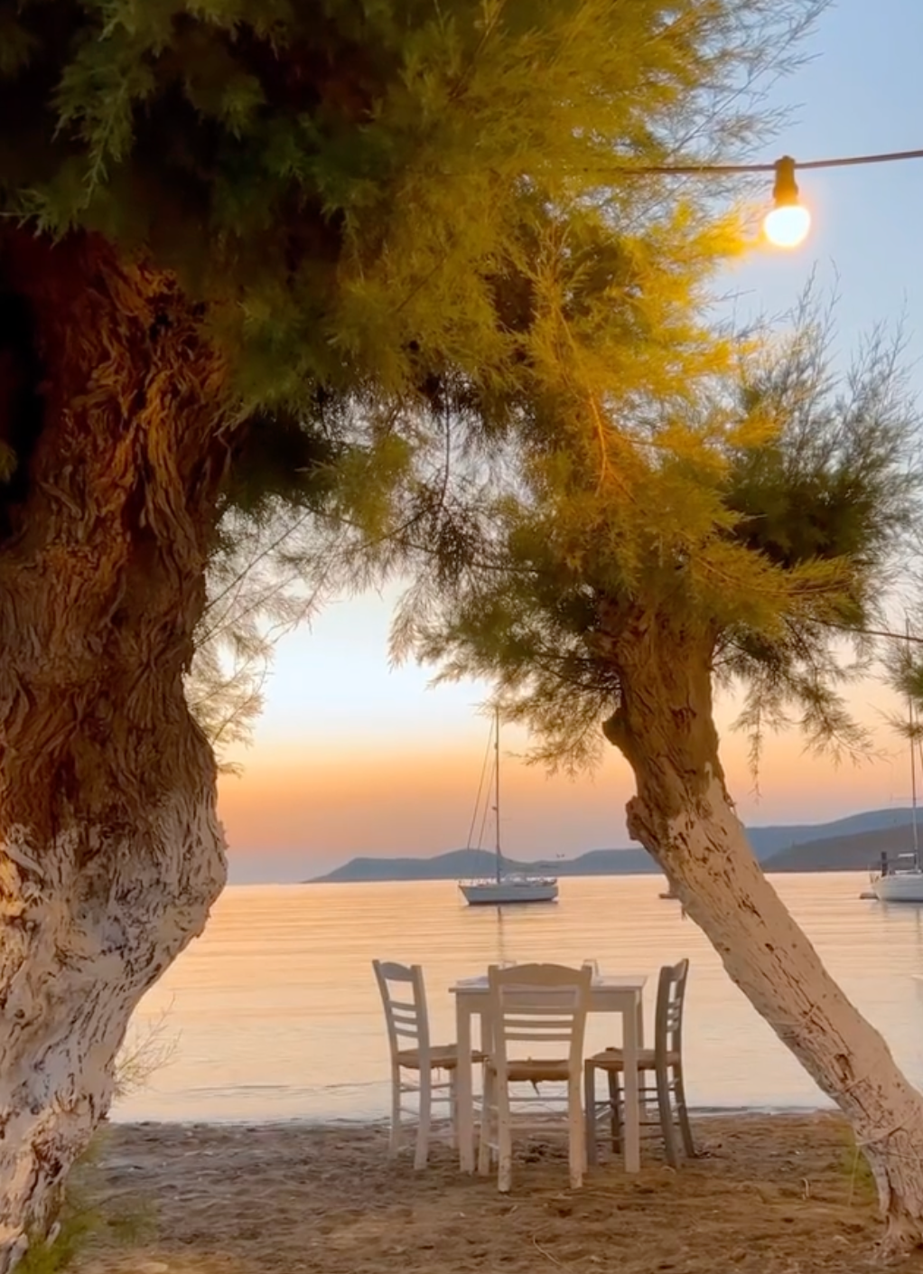 A white table with four chairs sits on a sandy beach beneath leafy trees at sunset. Sailboats float on calm water in the background, and a string light hangs above, creating a peaceful, inviting scene.