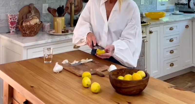 A person in a white blouse slices a lemon on a wooden cutting board in a bright kitchen. Lemons and garlic are on the table, with a bowl of lemons and various kitchen items in the background.