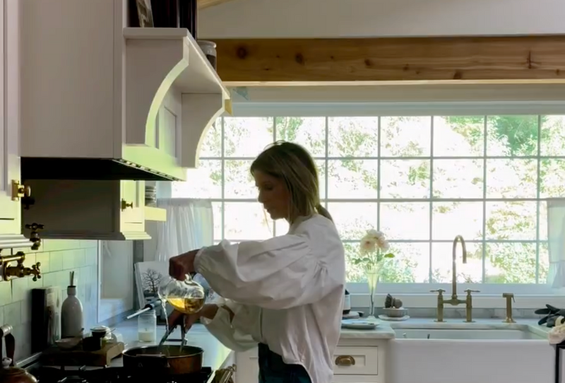 A woman in a white blouse stands in a bright kitchen, pouring liquid from a glass measuring cup into a pot on the stove. Large windows behind her let in natural light.