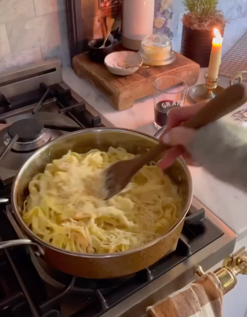 A hand stirs fettuccine pasta in a pan on a stove. Nearby are a candle, a glass of red wine, herbs, and kitchen items, creating a cozy cooking atmosphere.