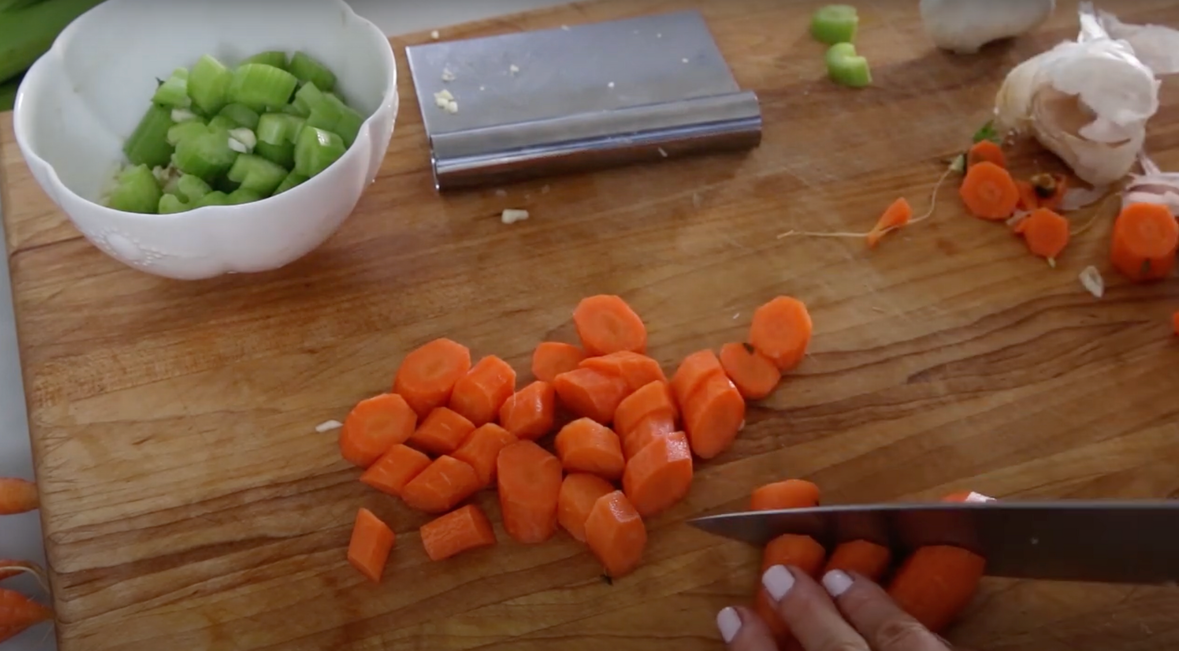 A hand slices carrots on a wooden cutting board with a knife. Chopped carrots are scattered on the board, and a bowl of chopped celery and a bench scraper are nearby. Garlic and celery pieces are also visible.