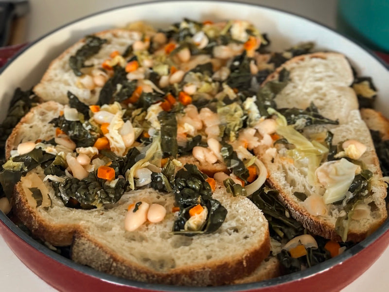 A casserole dish filled with slices of rustic bread layered with cooked greens, white beans, and diced carrots, creating a hearty vegetarian dish.