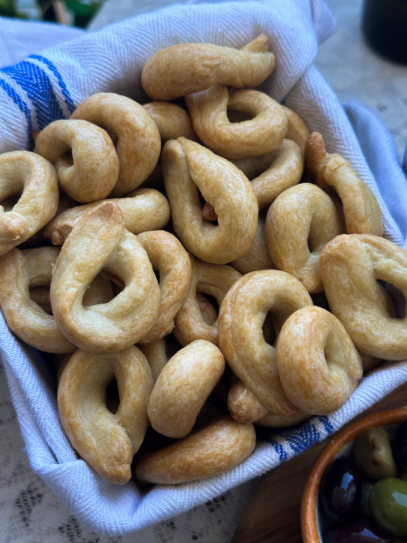 A basket lined with a white and blue cloth filled with ring-shaped Italian taralli crackers, with a bowl of olives partially visible at the bottom right corner.