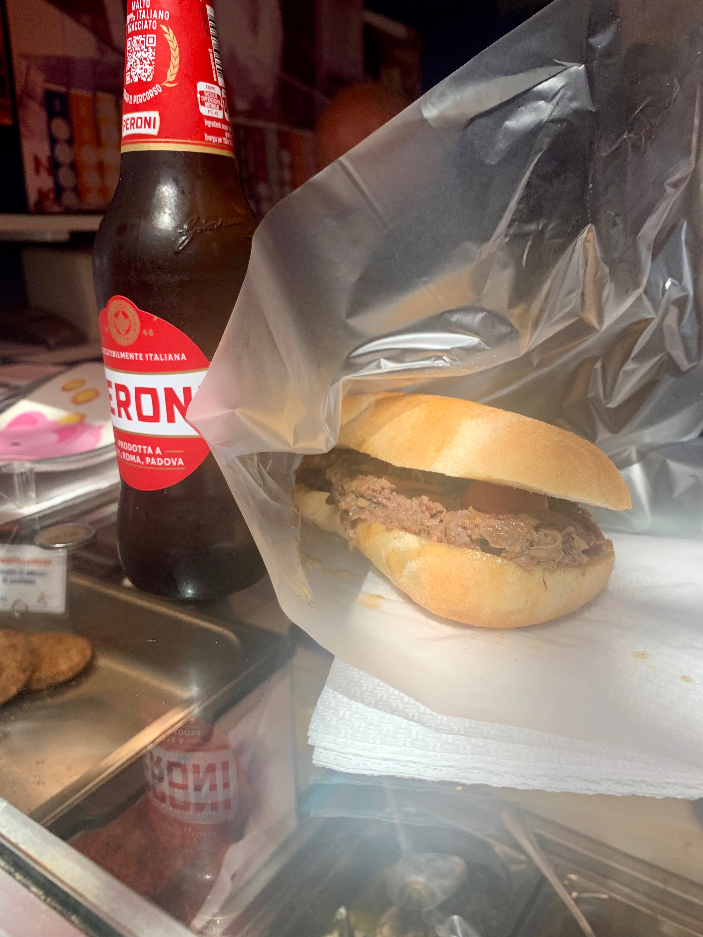 A sandwich filled with shredded meat sits on a napkin beside a bottle of Peroni beer. Both are placed on a glass counter, with a clear plastic sheet partially covering the sandwich.